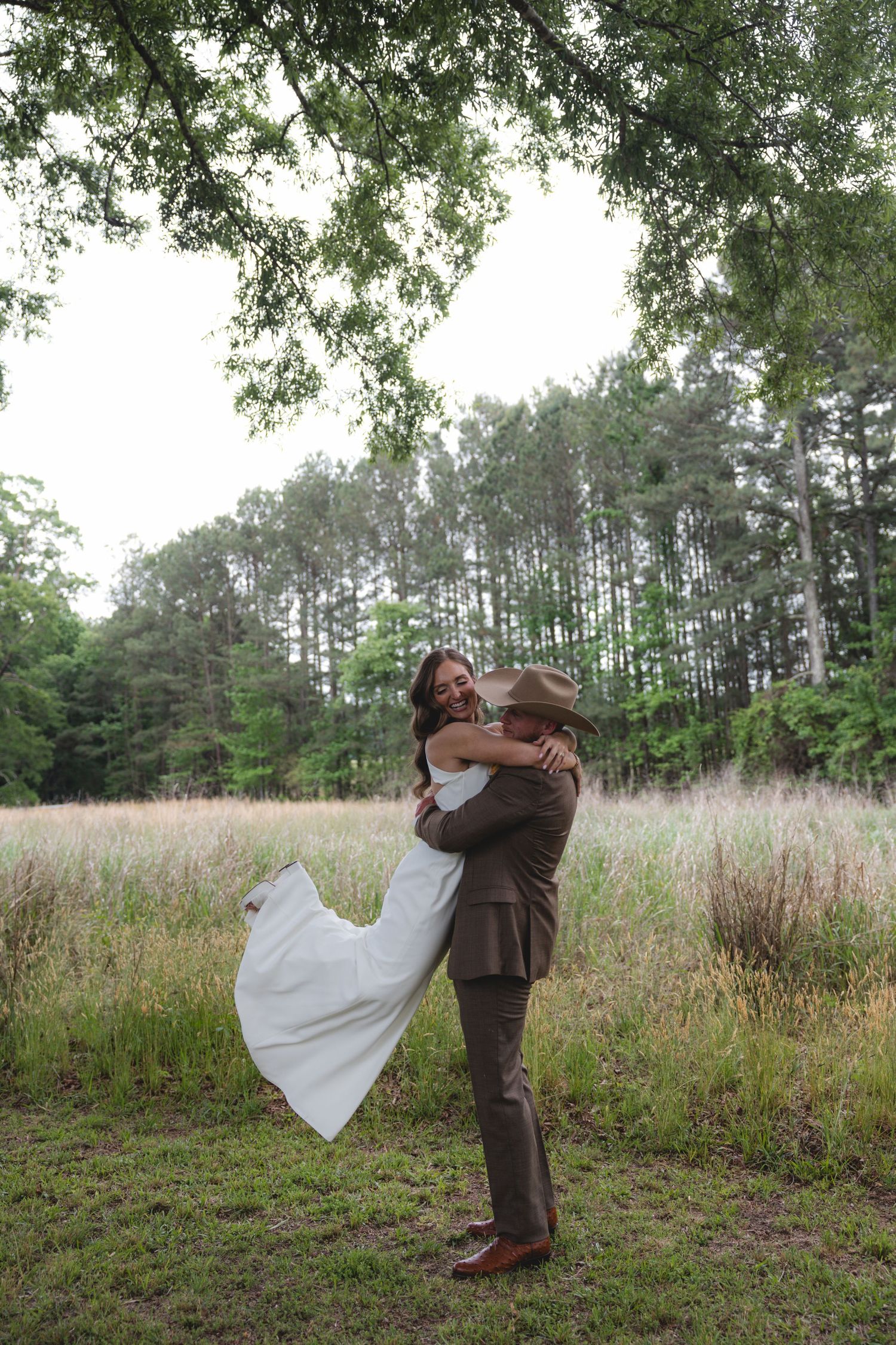 Groom in brown suit and cowboy hat lifting smiling bride in white dress in a lush green field.