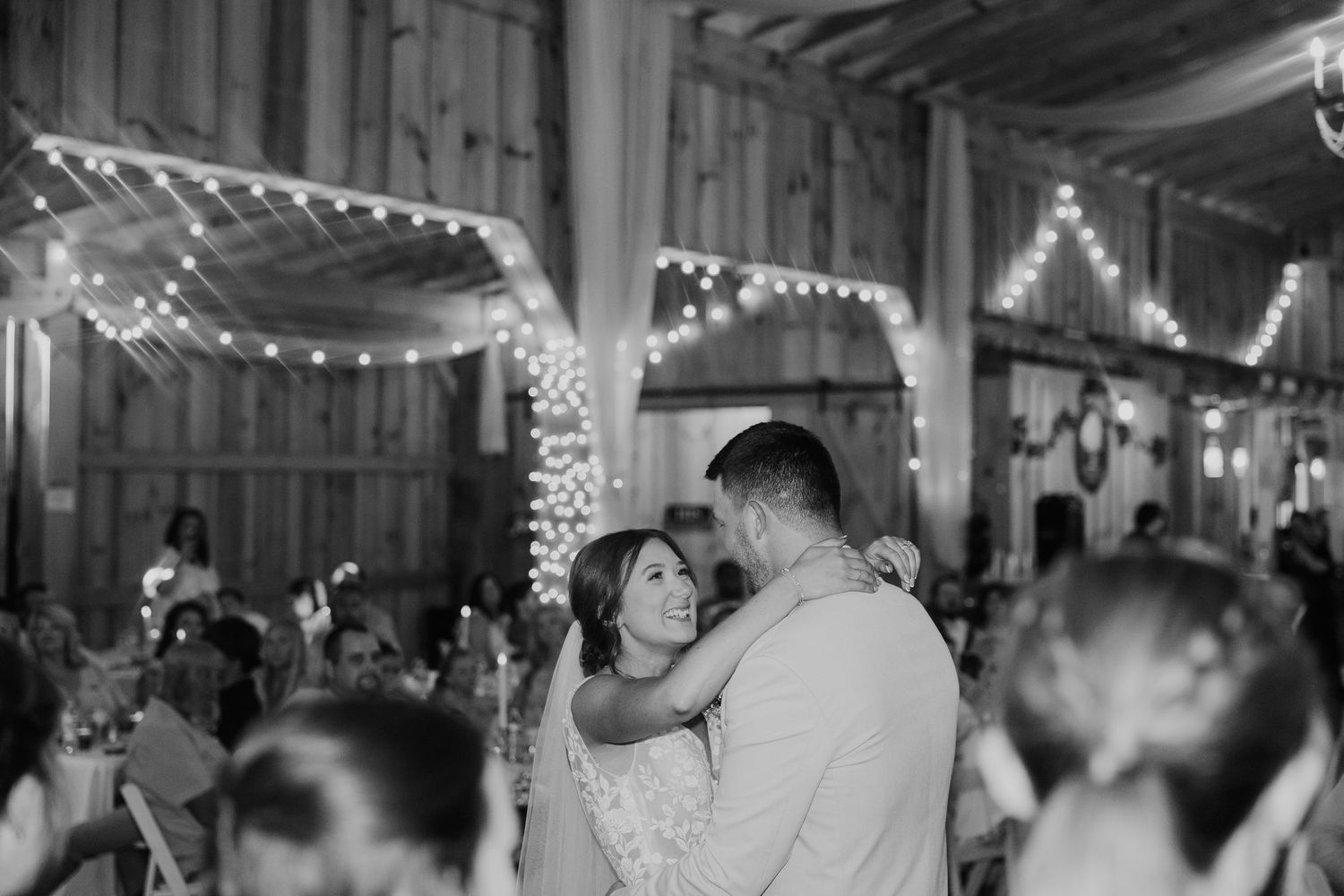 Black and white photo of bride and groom sharing first dance in a rustic barn with string lights.
