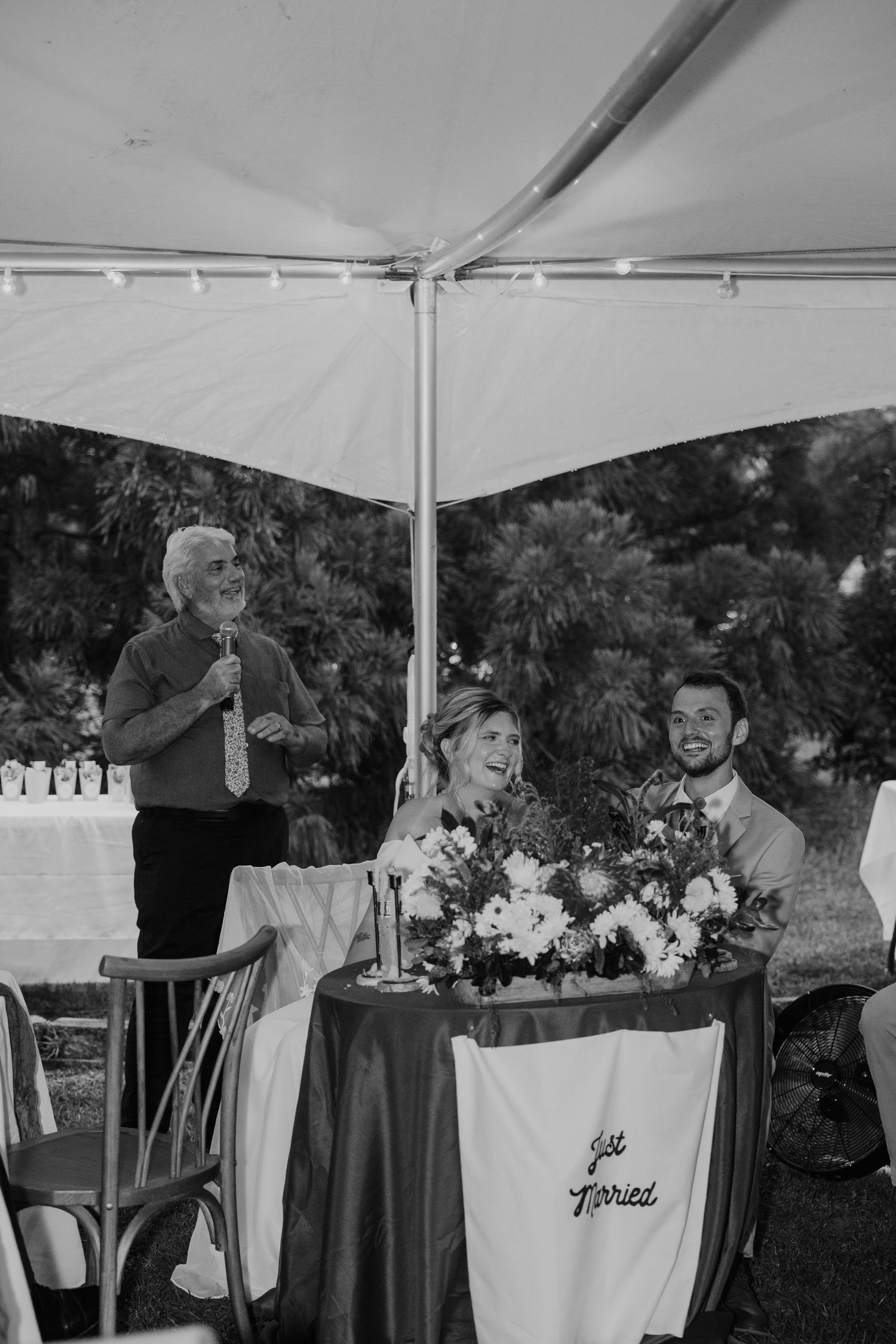 Black and white photo of newlyweds at sweetheart table with Just Married sign while man gives toast at outdoor wedding.