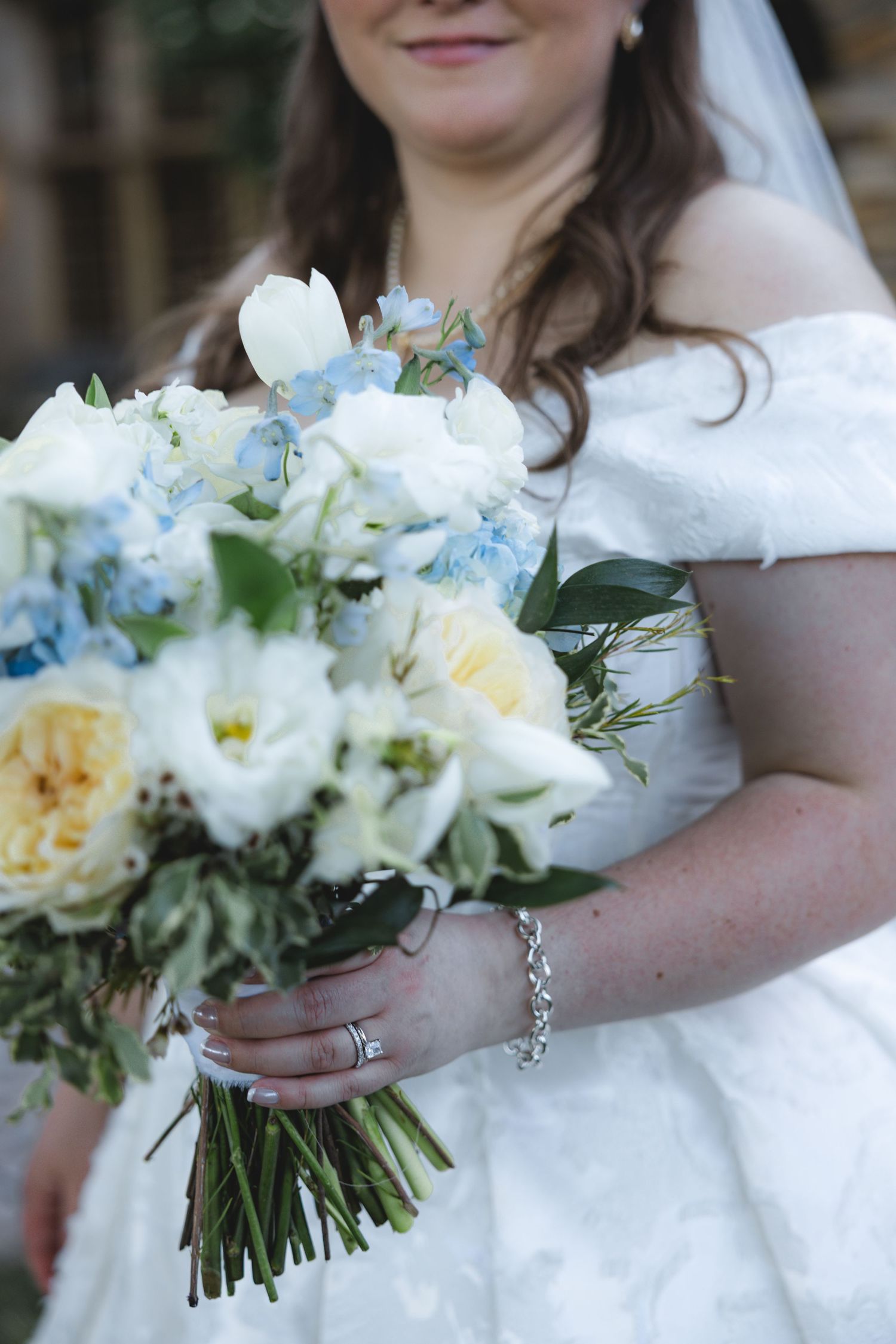 Bride in white off-shoulder gown holding a bouquet of blue, white, and yellow flowers with greenery.