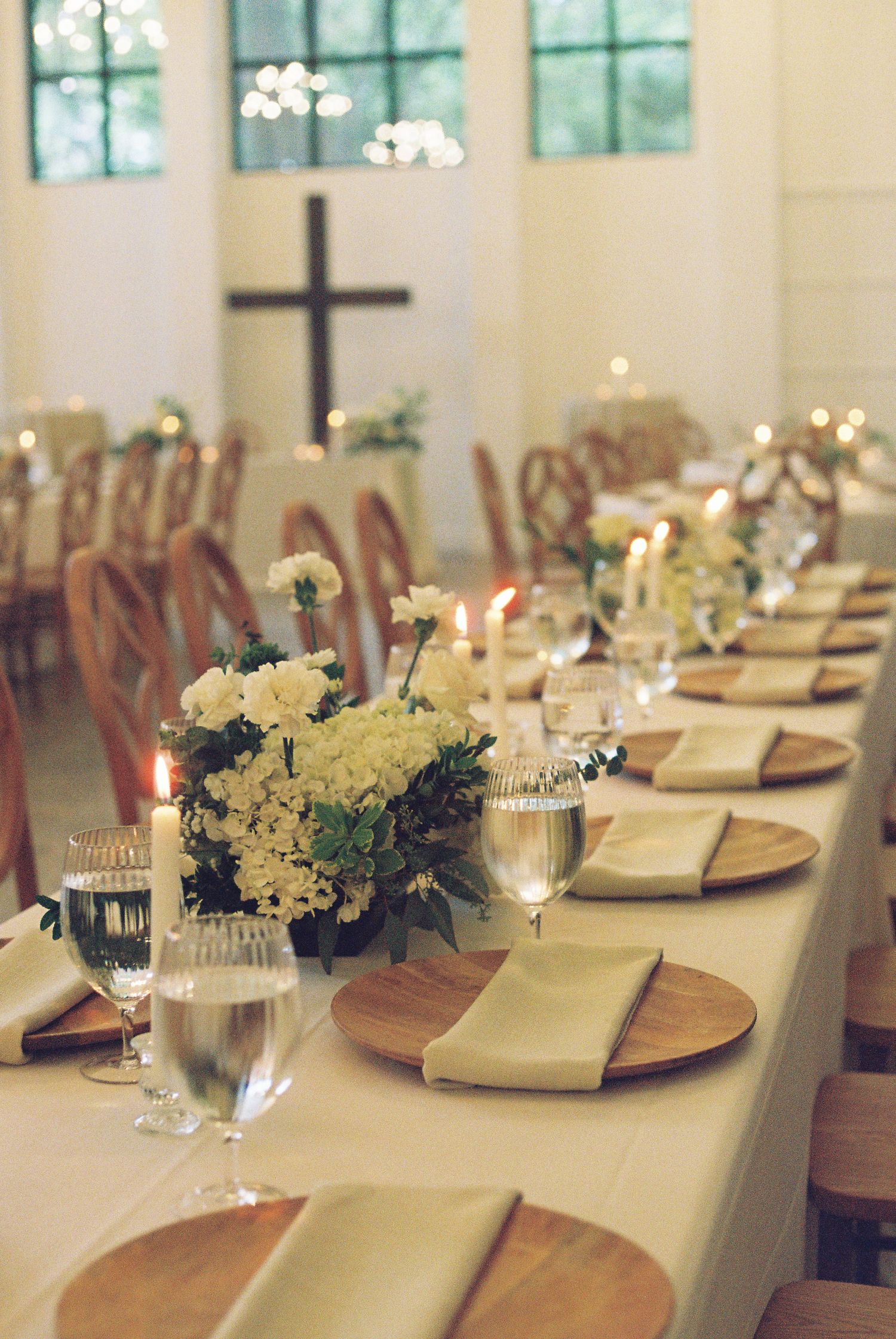 Elegant wedding reception table with wooden chargers, ivory napkins, white floral centerpieces, candles, and a cross in a white chapel.