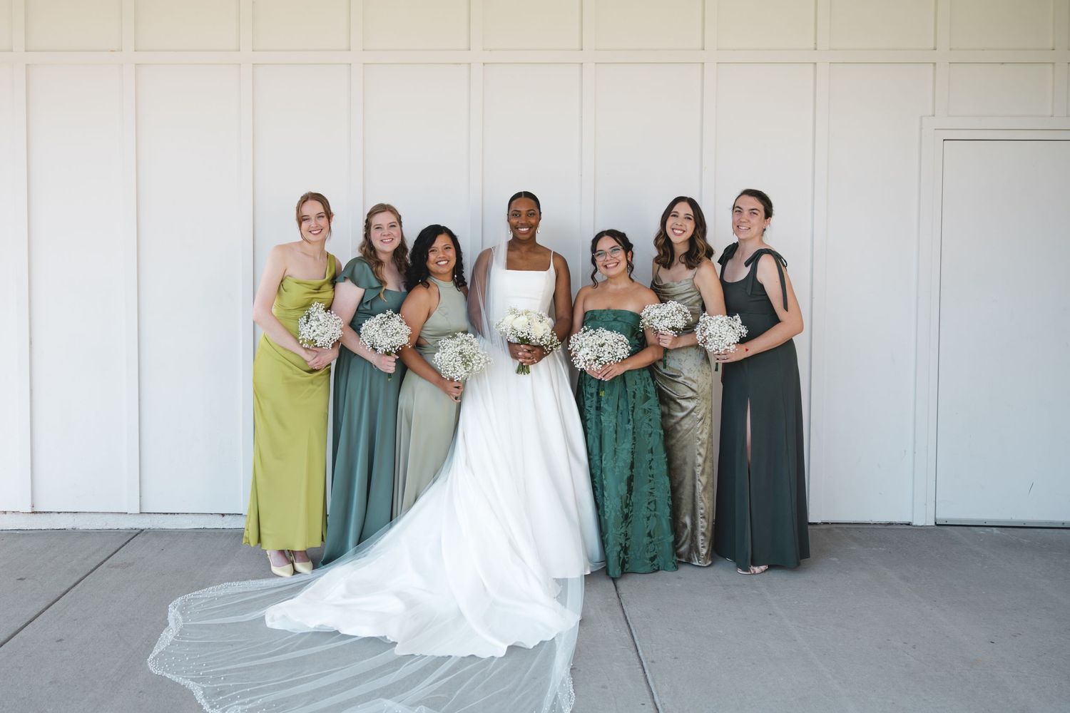 Bride in white gown with bridesmaids in green dresses holding white bouquets, posing outdoors against a white wall.
