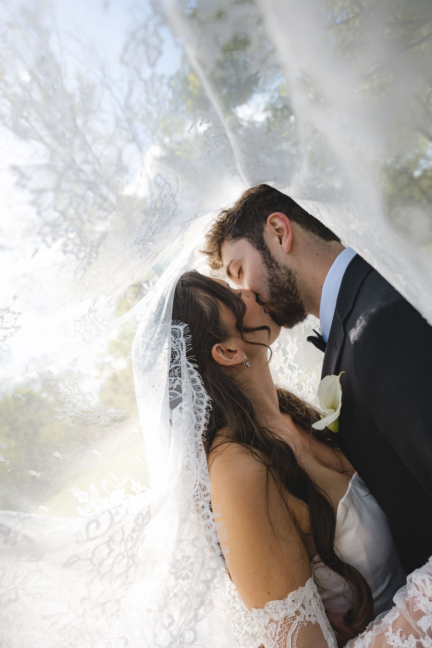 Bride and groom sharing a romantic kiss under a flowing lace wedding veil outdoors.