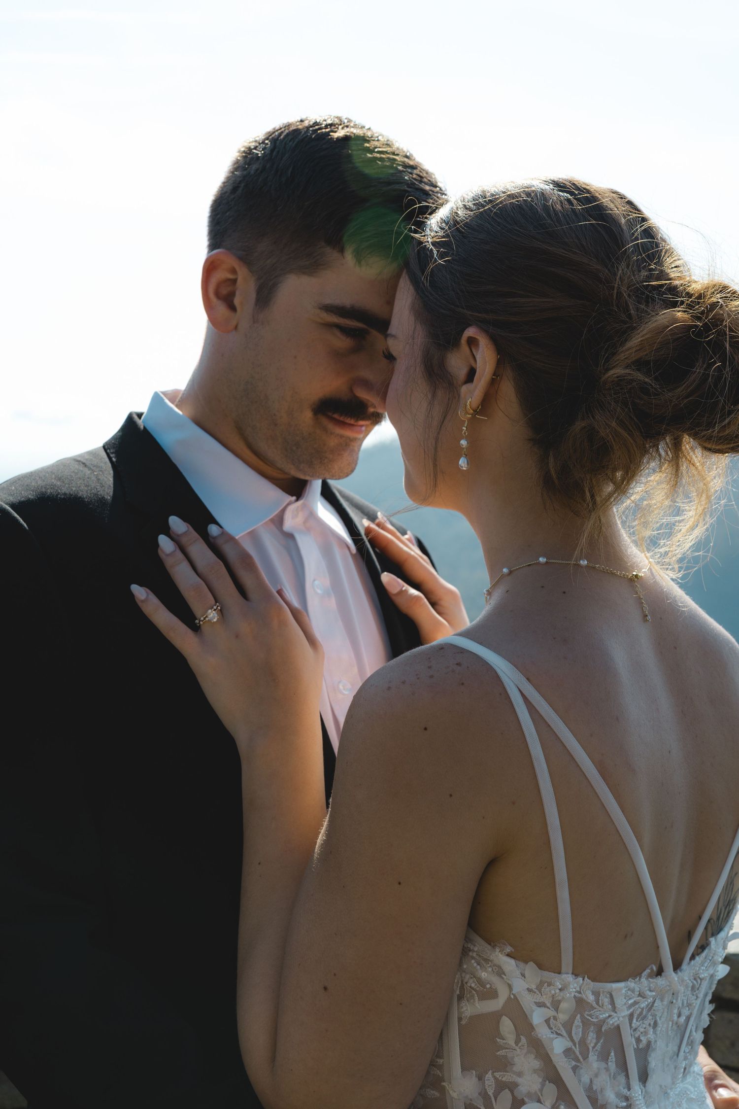 Bride and groom share an intimate moment, touching foreheads outdoors, bride in lace gown with diamond earrings.