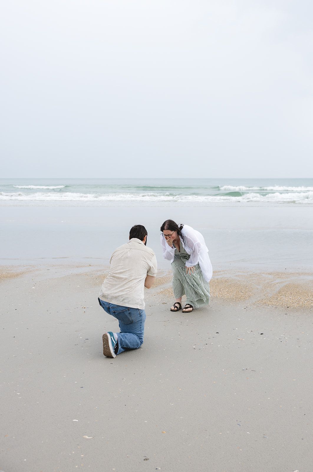 Man kneeling on beach proposing to emotional woman in white shirt and green skirt by ocean waves.