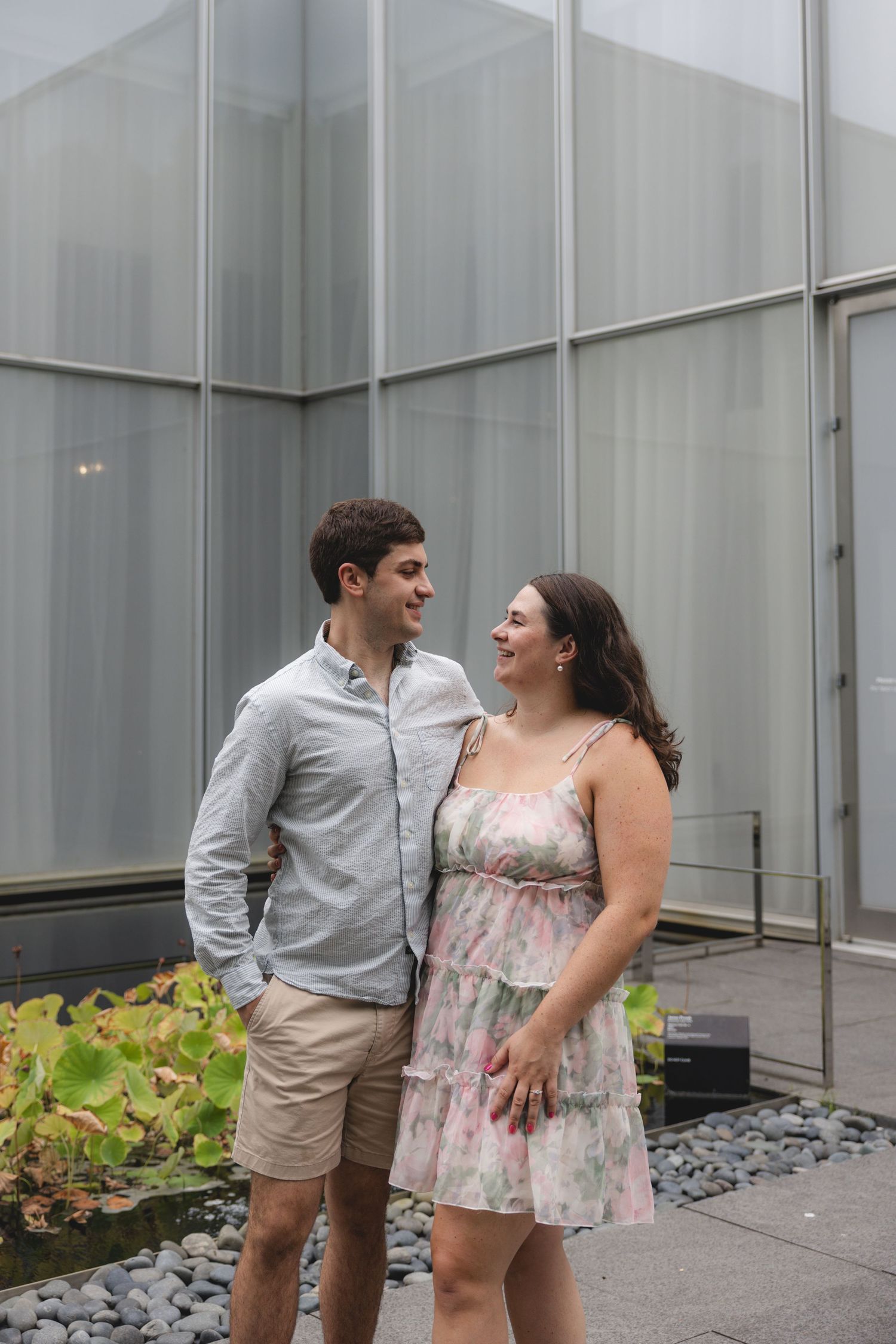 A smiling couple stands together outdoors near a modern glass building, man in gray shirt and shorts, woman in floral dress.
