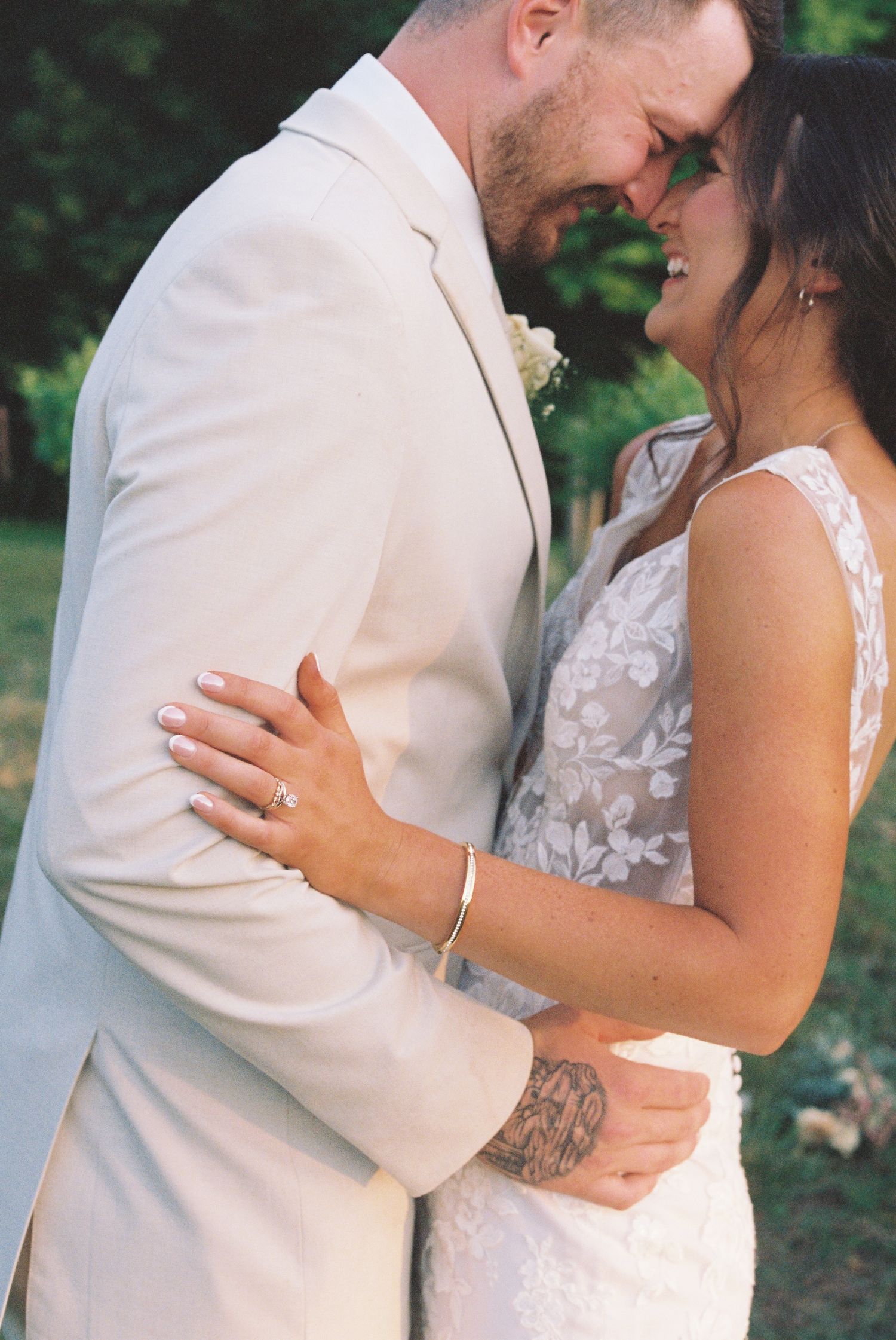 Bride and groom embracing tenderly in outdoor wedding attire, bride wearing lace dress and gold bracelet.