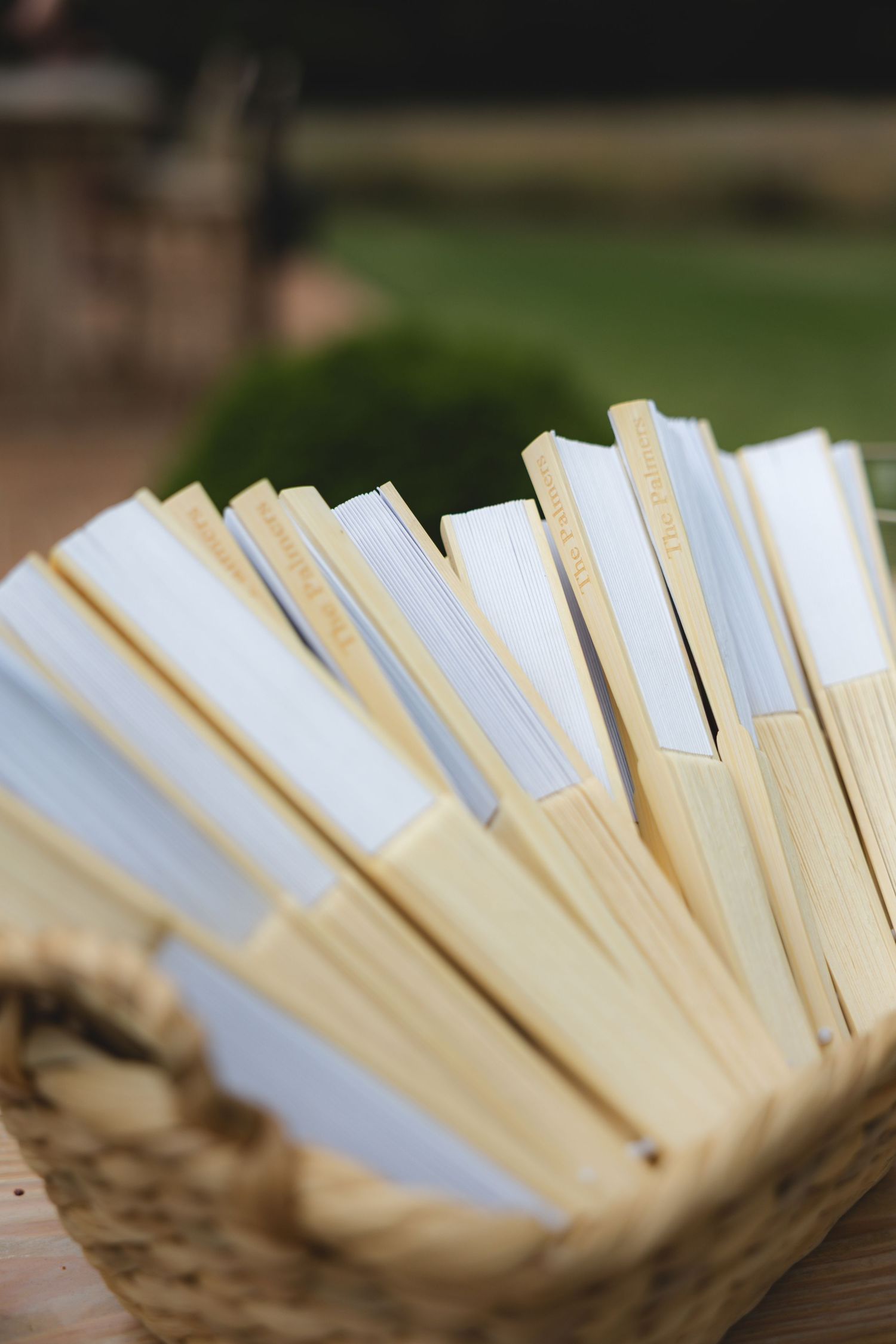 Wooden hand fans with white paper programs arranged in wicker basket for outdoor wedding guests.