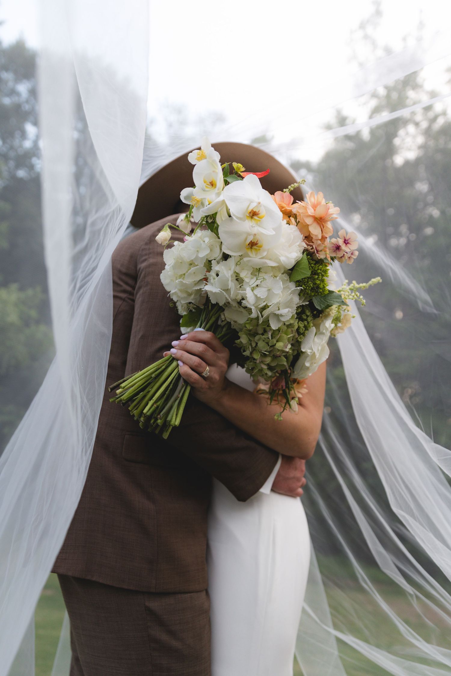 Bride and groom embracing under a flowing veil, bride holding a lush white and peach floral bouquet.