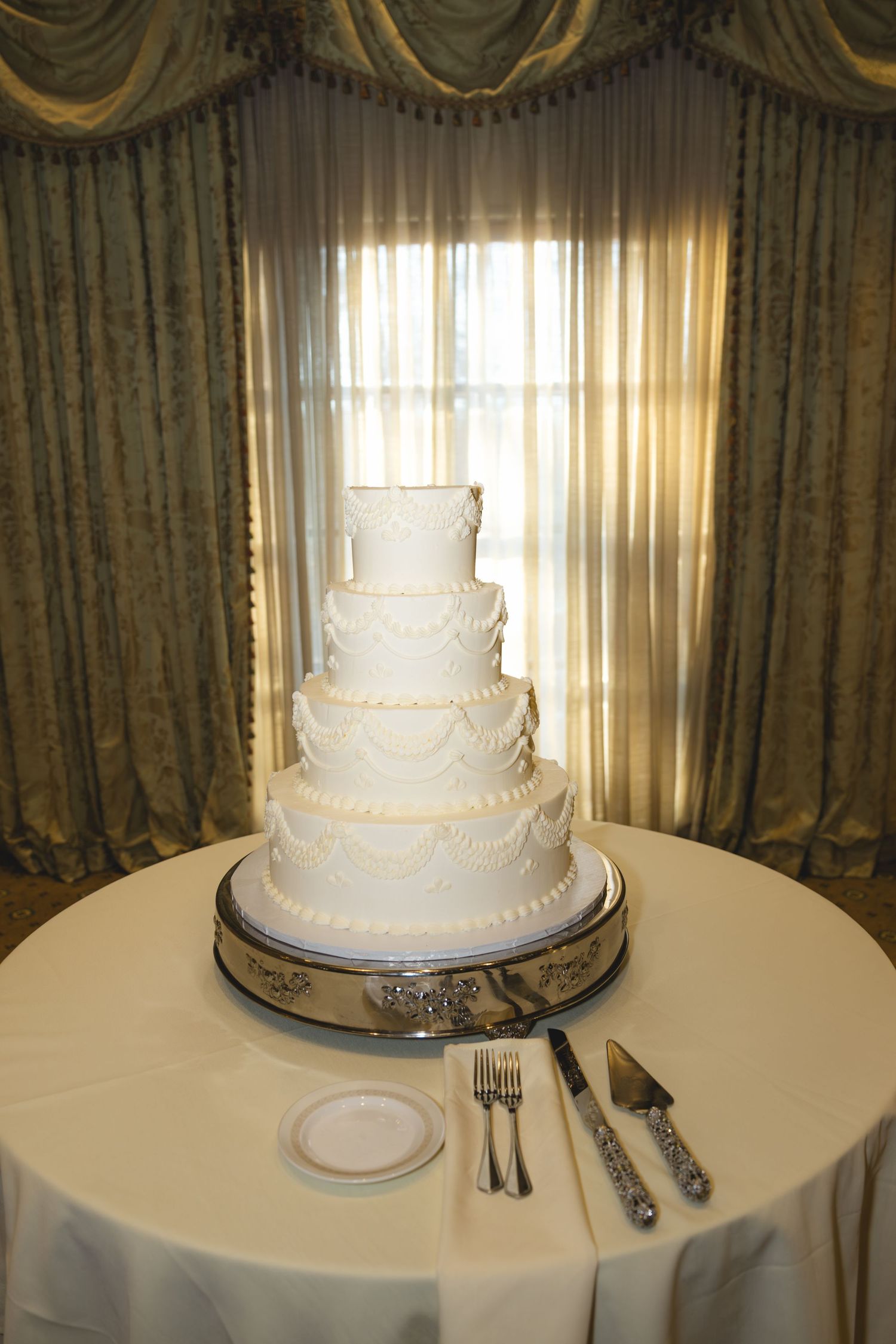 Elegant four-tier white wedding cake with draped fondant details on silver stand before draped window curtains.