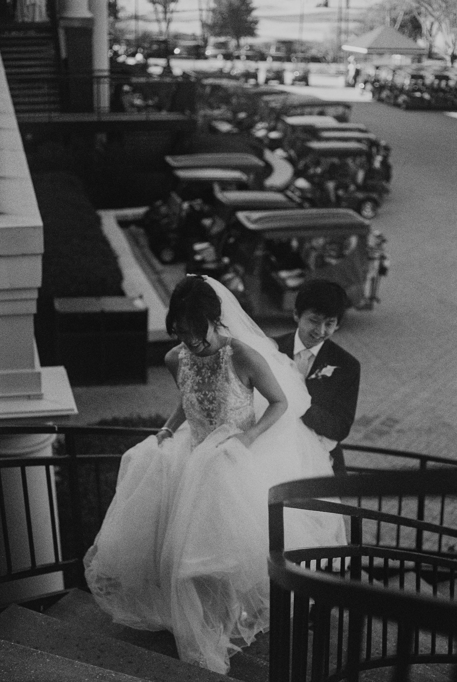 Black and white photo of bride in lace gown and groom descending outdoor staircase at wedding venue.