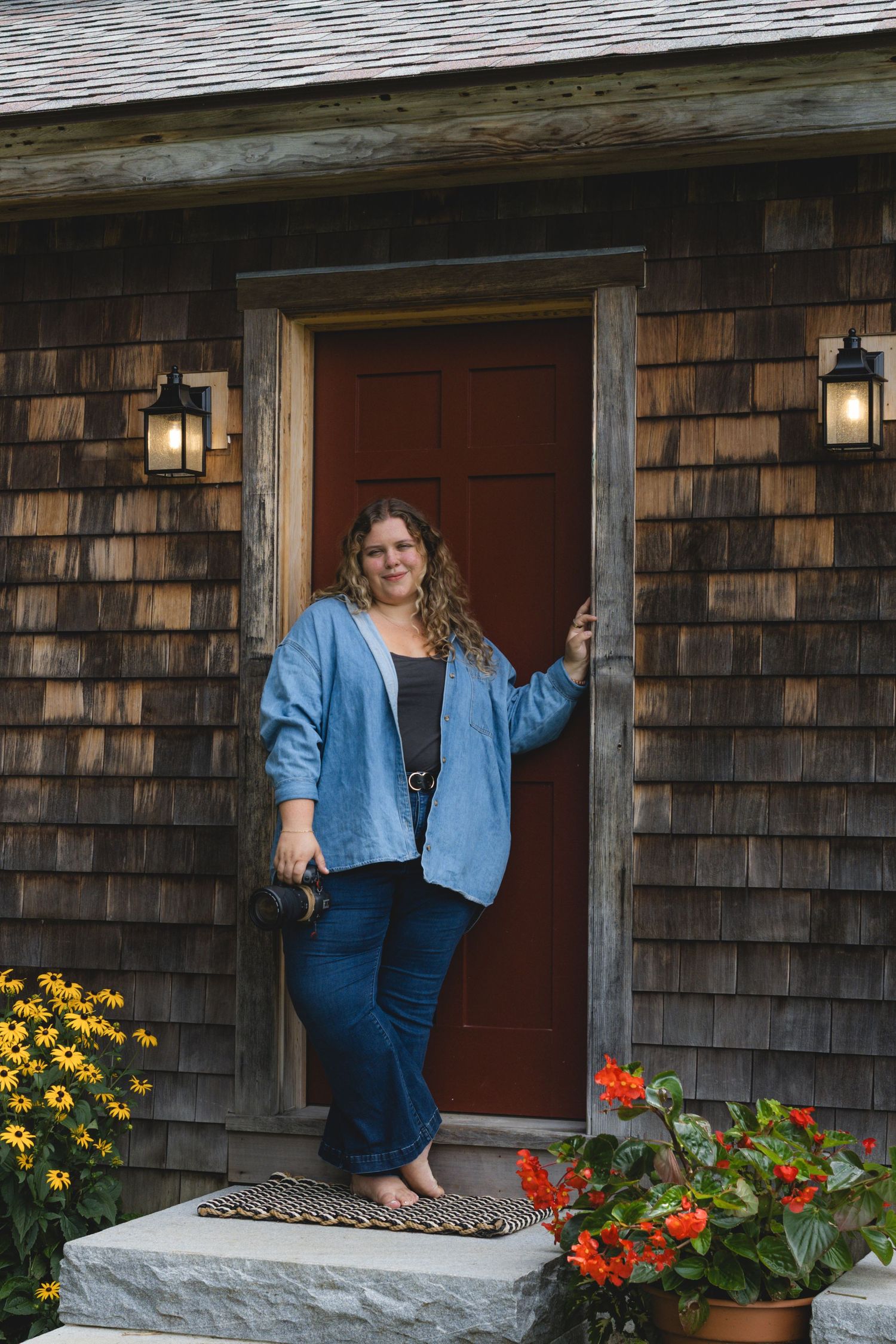 Woman in blue jacket and jeans posing at red door of cedar shingle home with flower pots.