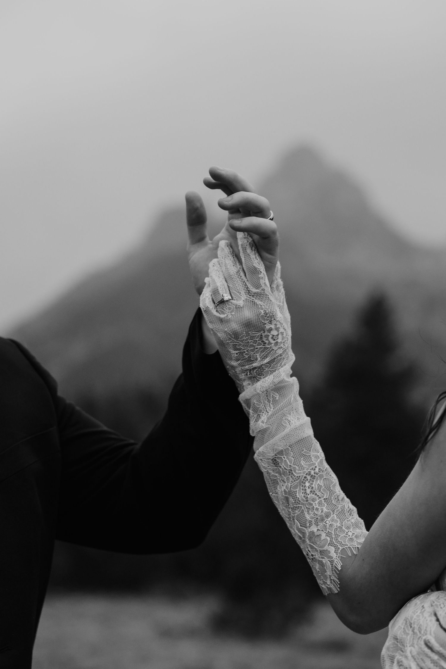 Black and white photo of bride in lace gloves holding groom's hand with mountain backdrop.