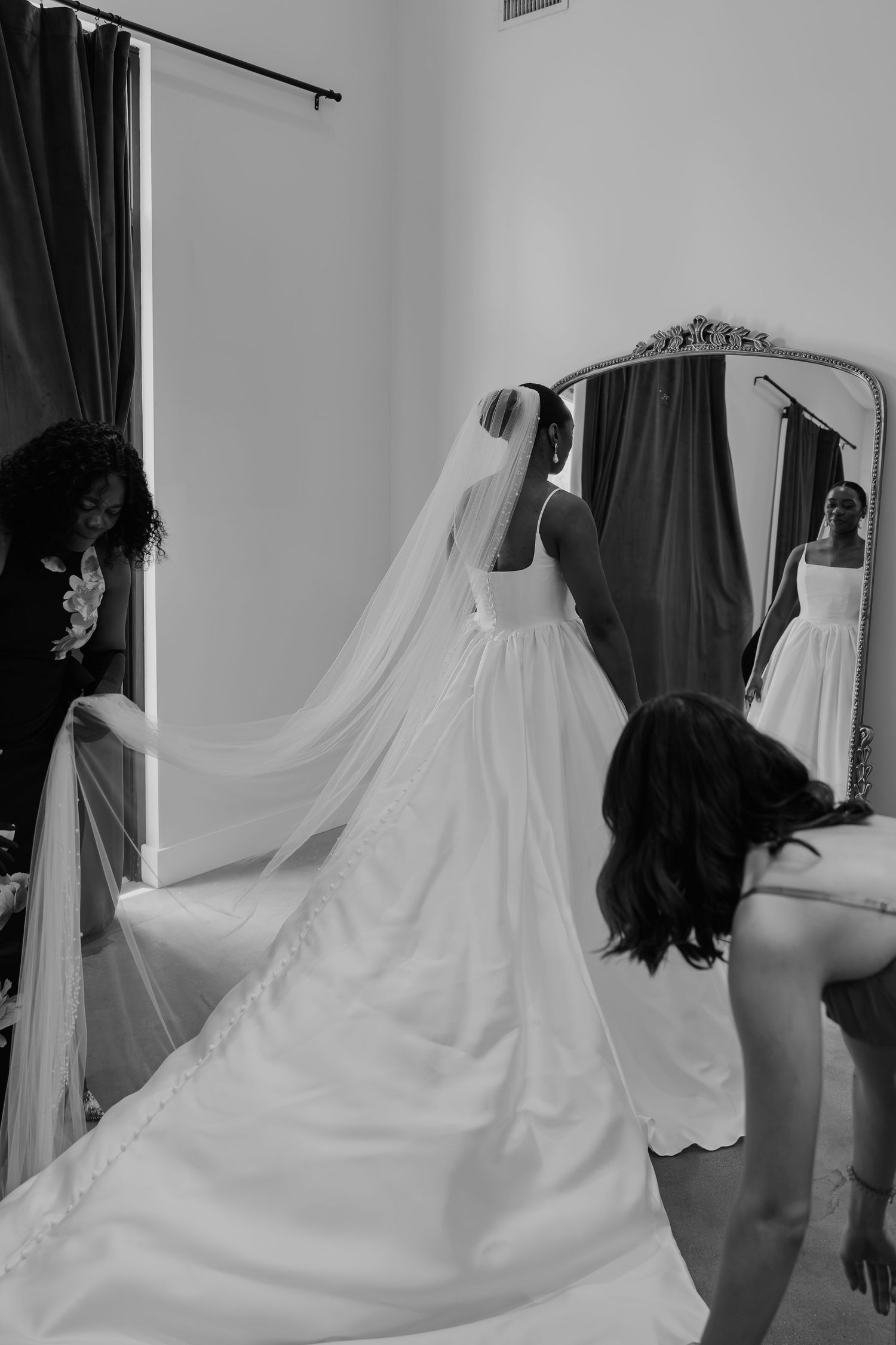 Black and white photo of a bride in a wedding gown and veil being assisted by two women near an ornate mirror.