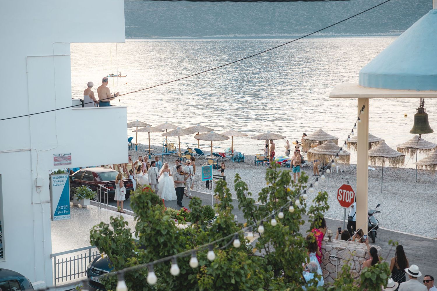 Aerial view of a coastal beach town with tourists, umbrellas, and a waterfront promenade on a sunny day.