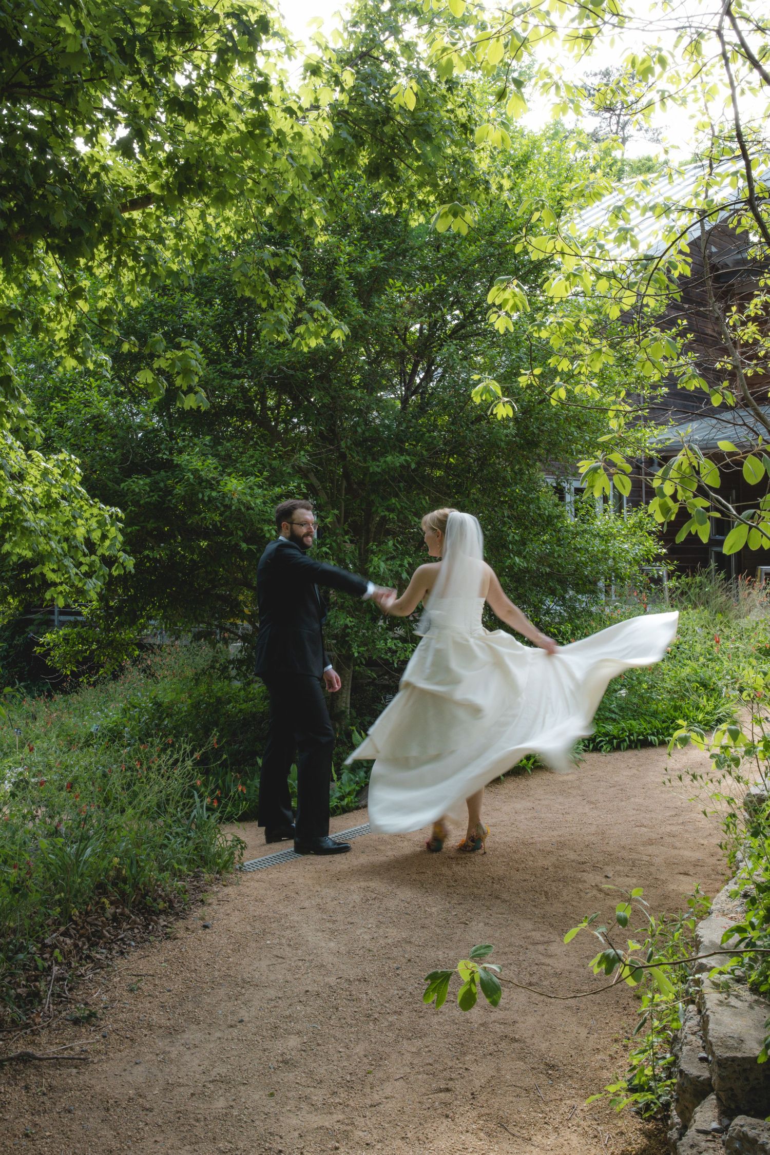 Bride in flowing white gown twirls with groom on garden path surrounded by lush green trees.
