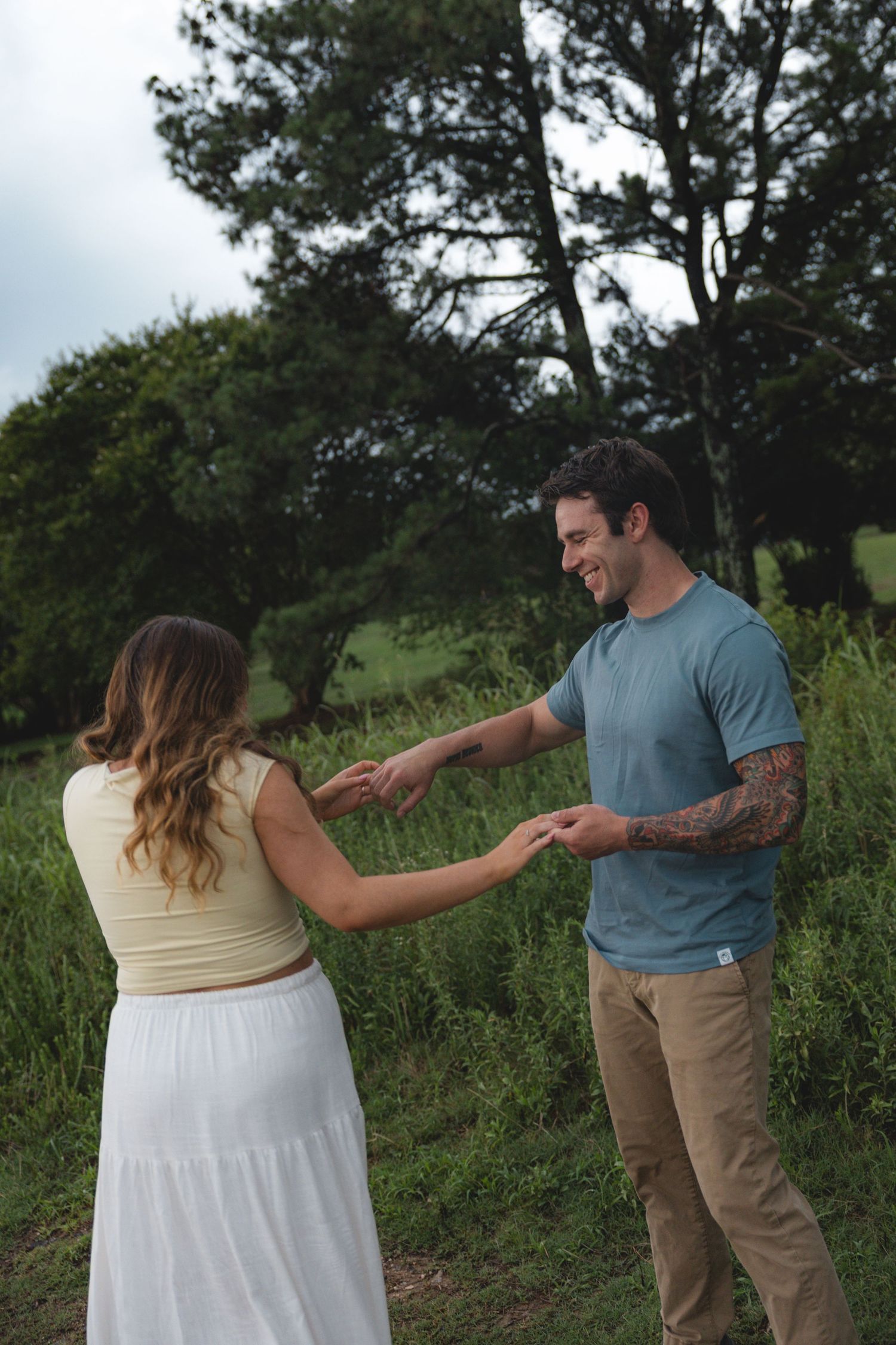 A smiling couple holds hands in a lush green field, man with tattoos wearing blue tee, woman in white skirt.