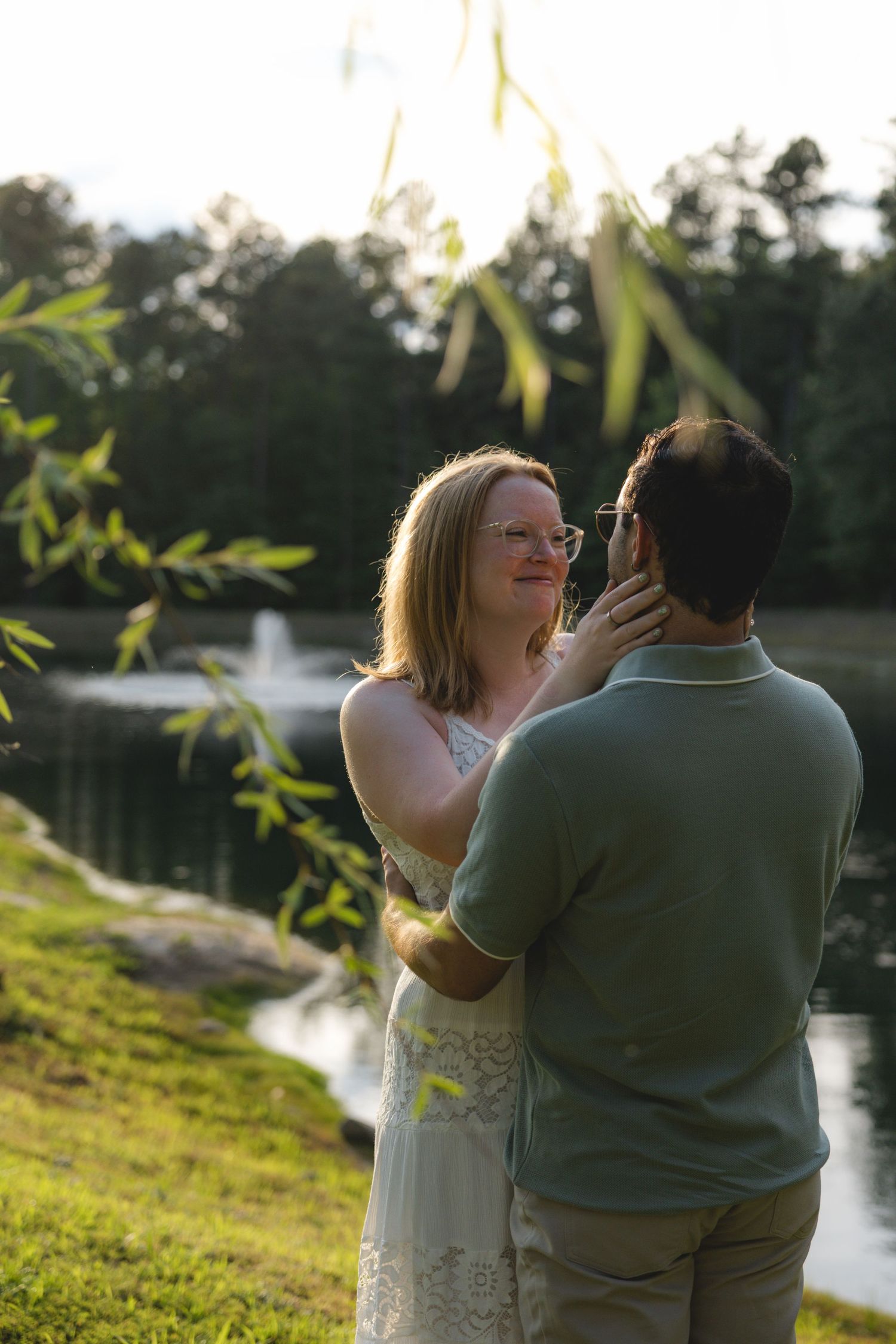 Couple sharing a romantic moment by a pond with a fountain, surrounded by lush greenery at golden hour.