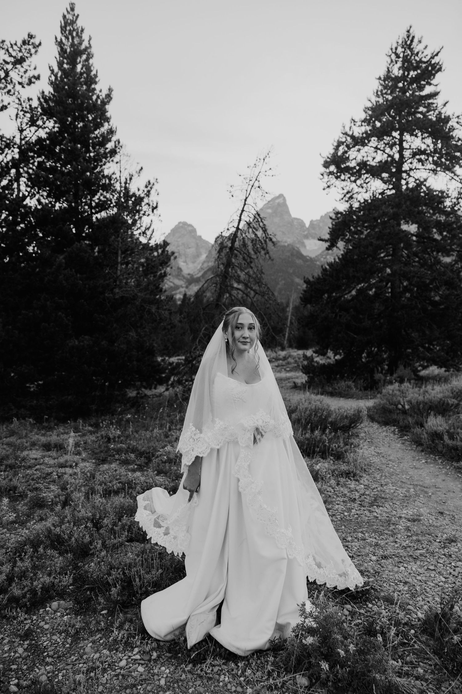 Black and white photo of a smiling bride in a lace veil and gown posing in a mountain forest setting.