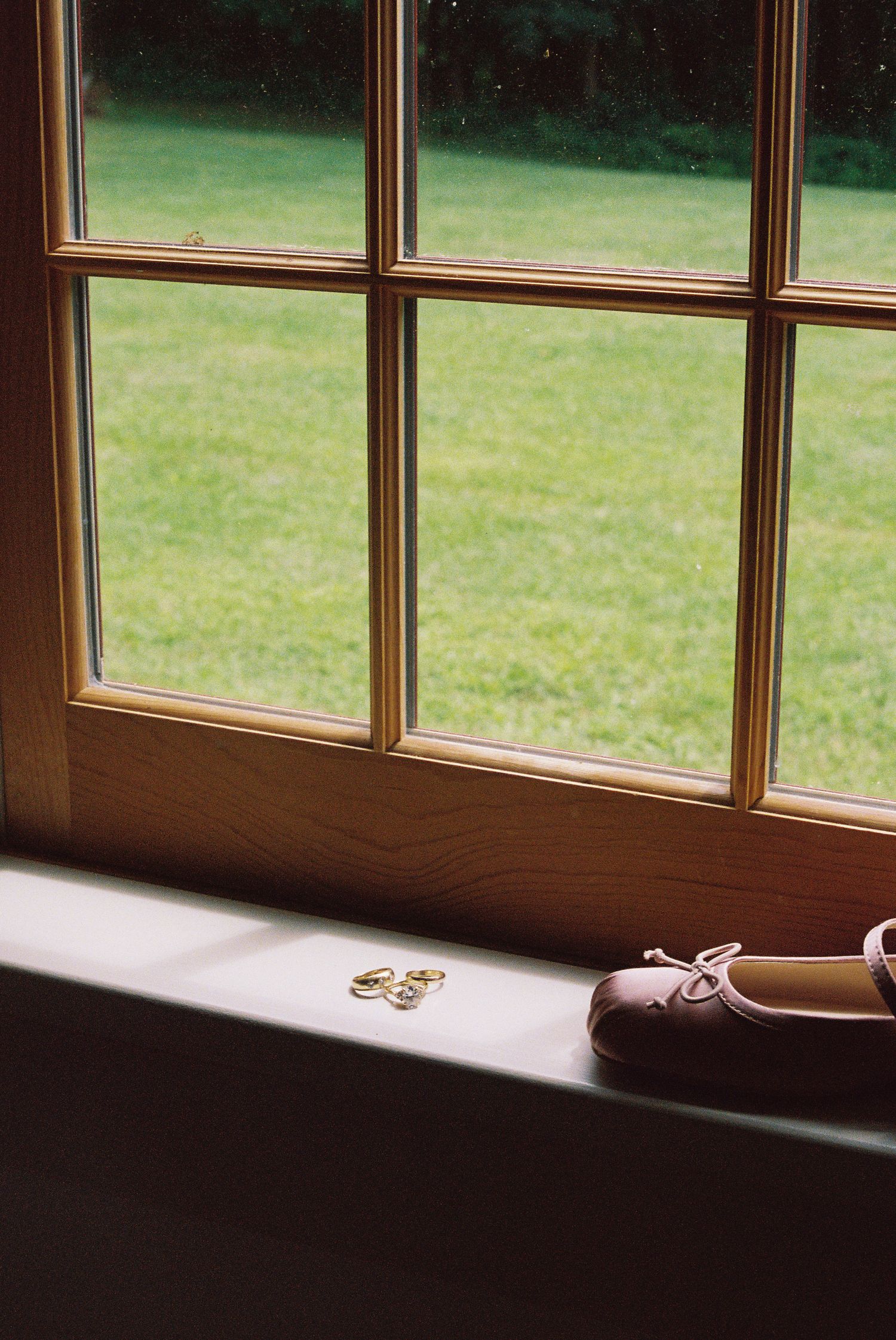 Wedding rings and bridal ballet flats on a sunlit wooden windowsill overlooking a lush green lawn.