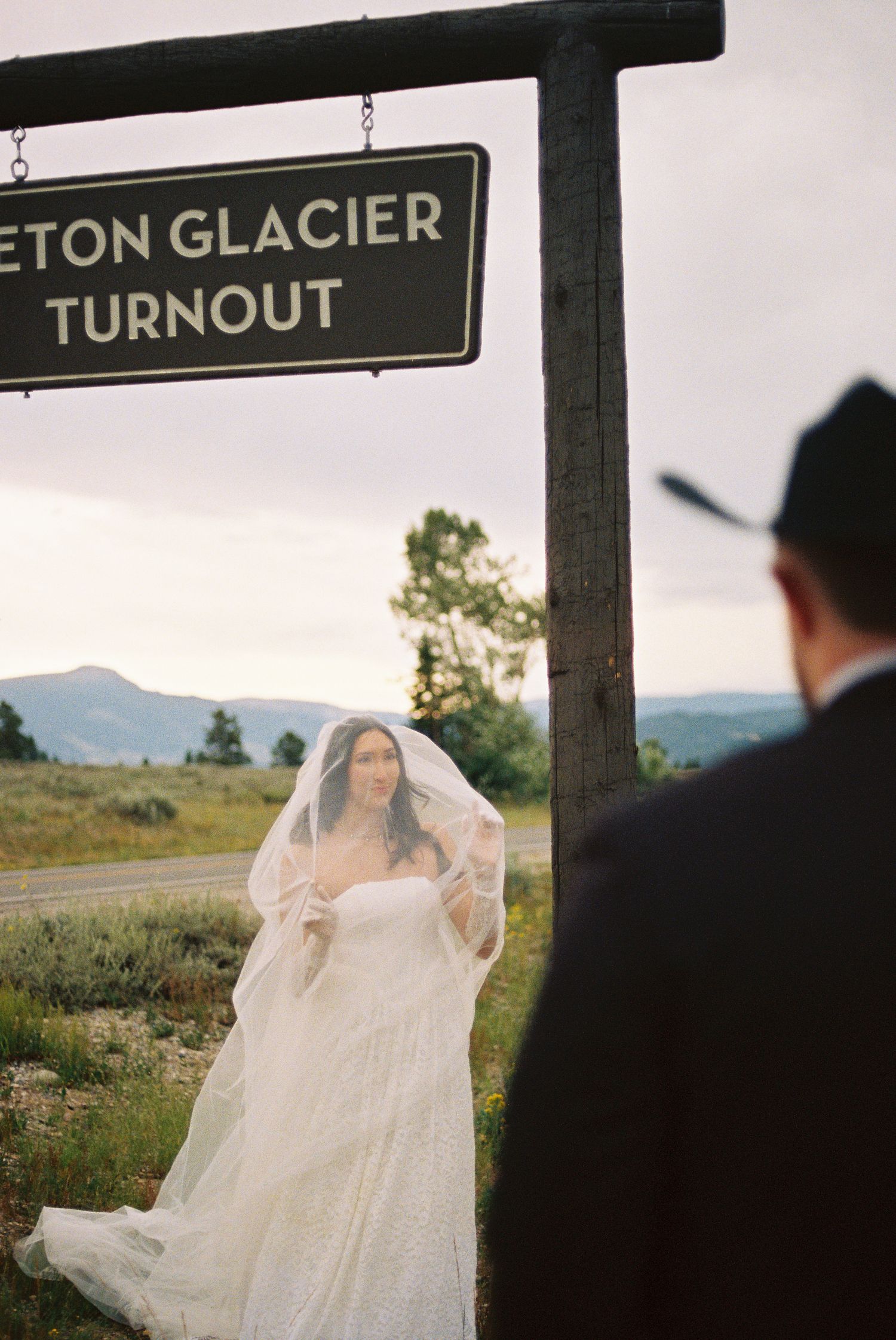 Bride in white gown and veil stands beneath Teton Glacier Turnout sign in Grand Teton National Park.