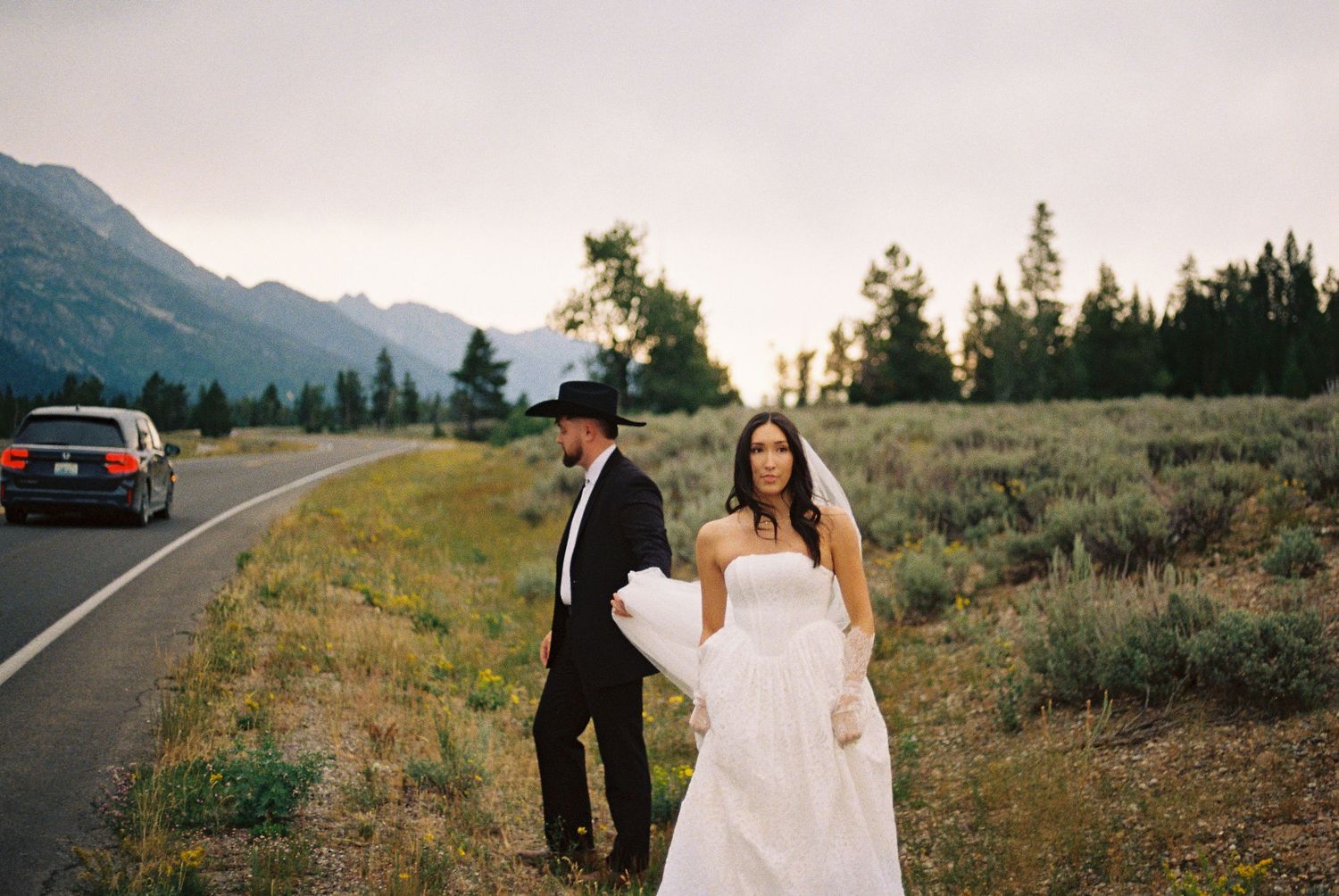 Bride in white gown and groom in black suit with cowboy hat pose on mountain road at sunset.