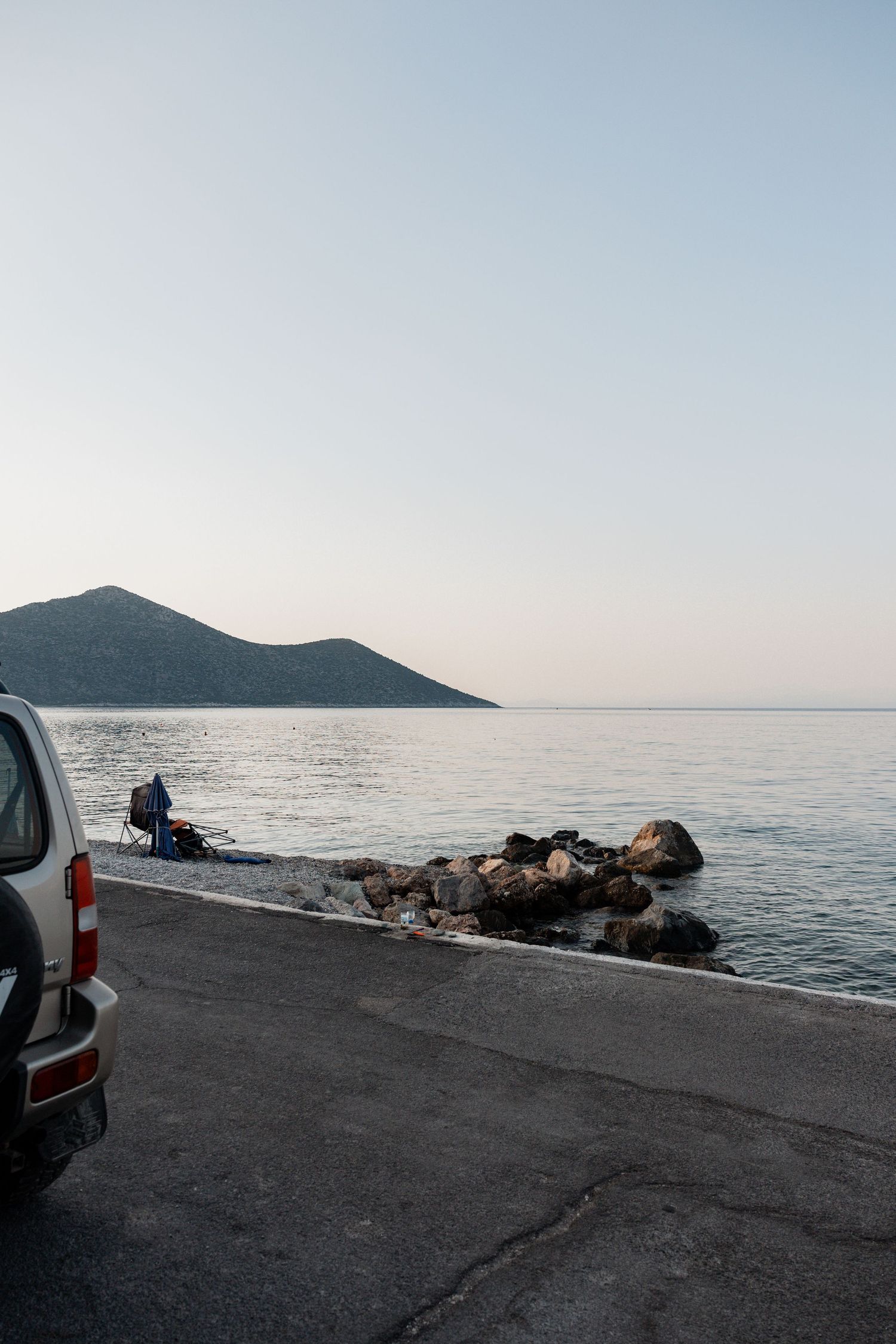 Person fishing by rocky seaside road at dusk with a mountain silhouette and calm sea in the background.