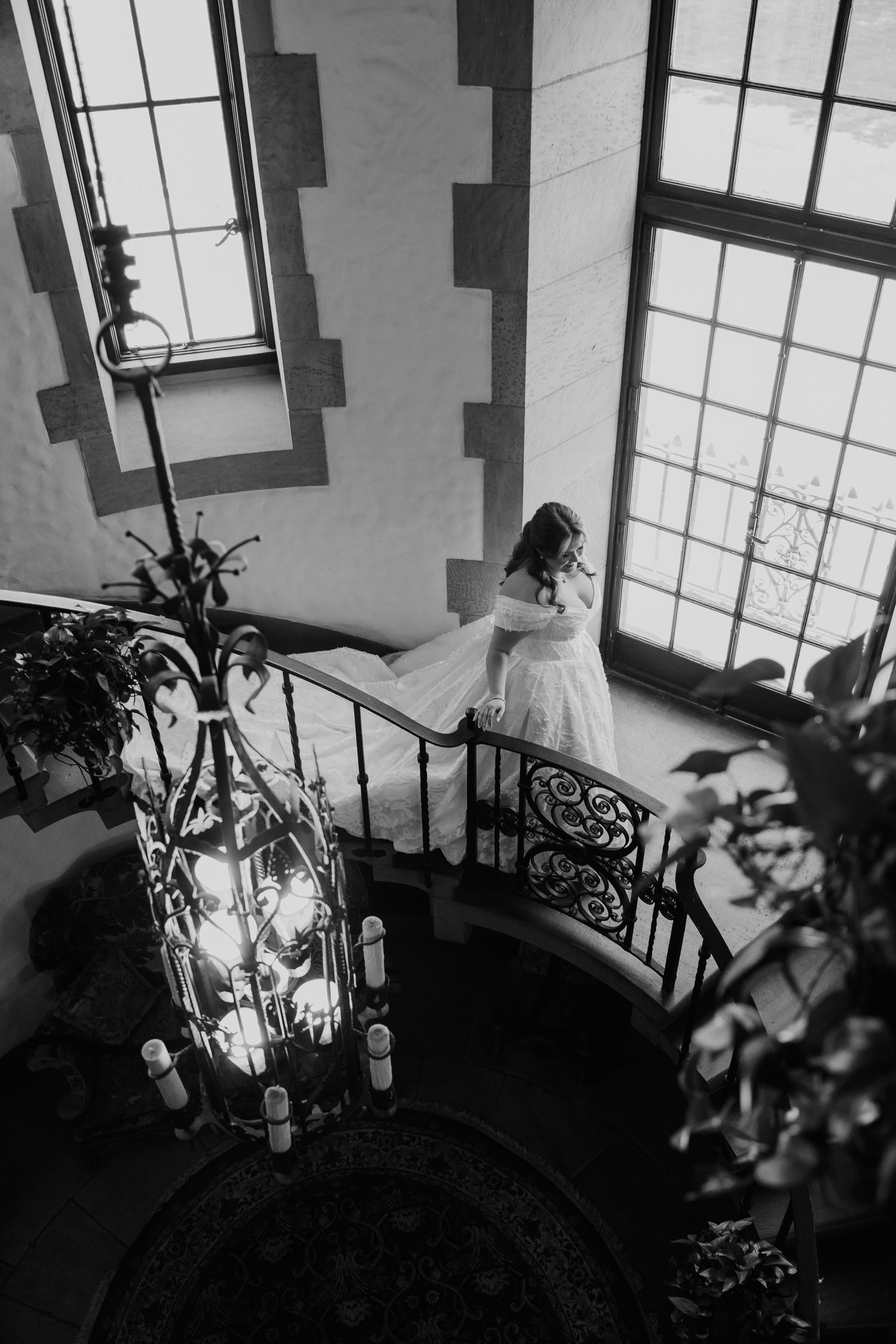 Black and white photo of a bride in a wedding gown descending an elegant spiral staircase with wrought iron railing.