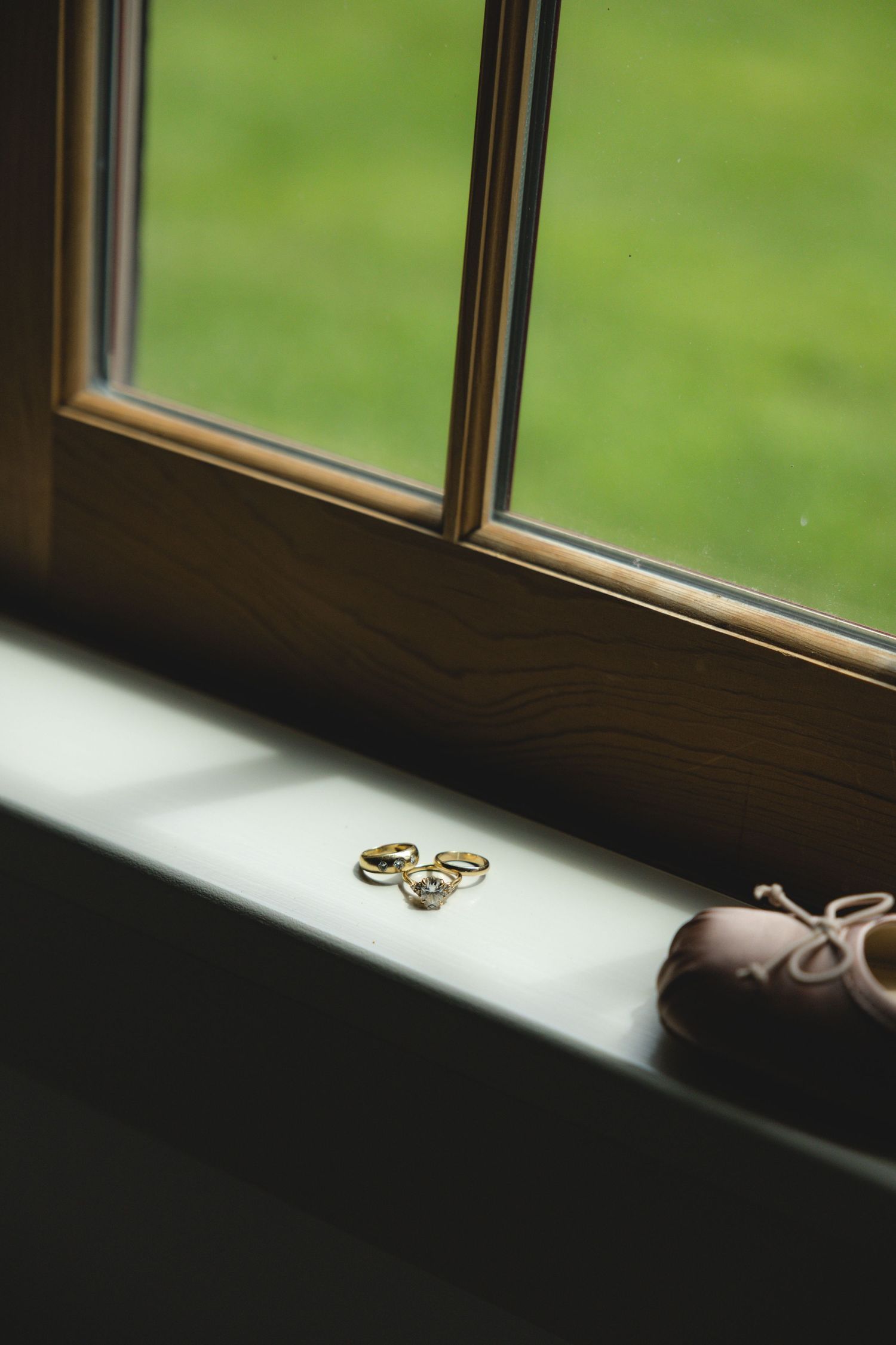 Gold wedding rings and an engagement ring rest on a sunlit windowsill beside blush bridal shoes.