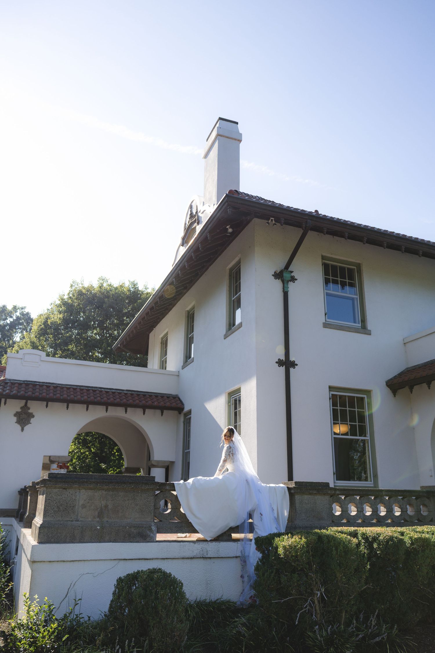 Bride in white gown posing on stone terrace outside elegant white stucco mansion with Spanish-style roof on sunny day.