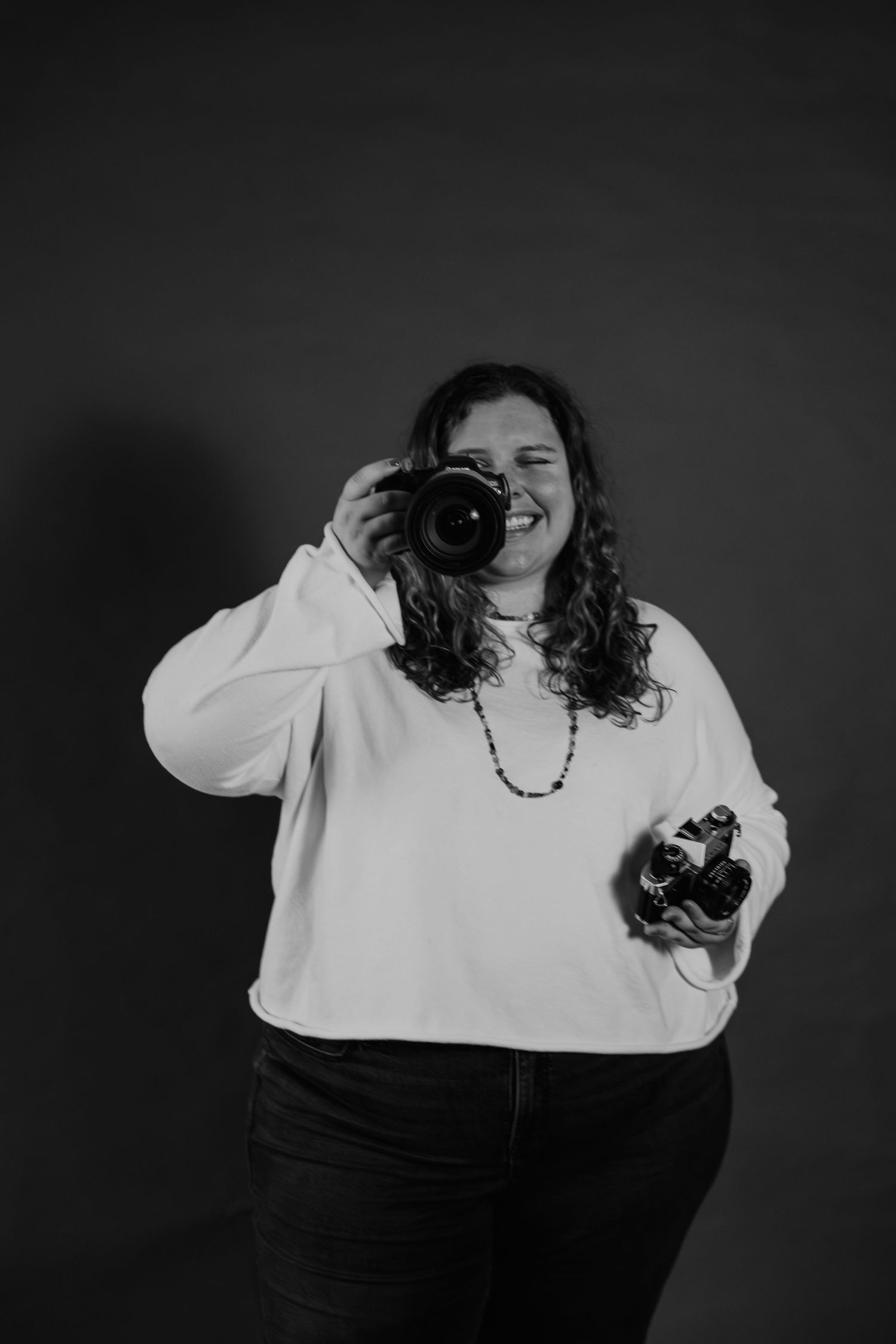 Black and white photo of a smiling woman holding two cameras, wearing a white sweatshirt and beaded necklace.