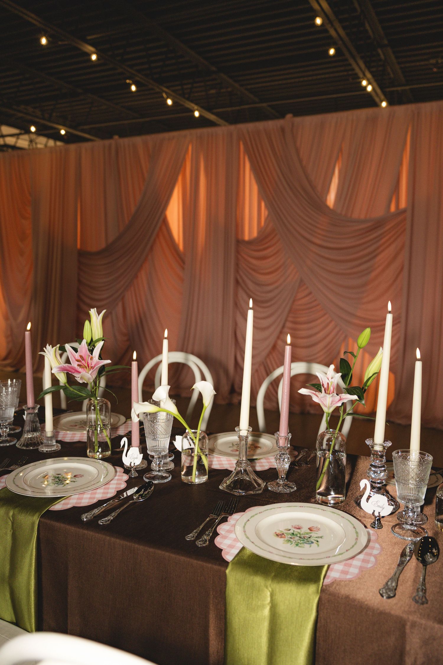 Elegant wedding reception table with green runners, pink lilies, tall candles, and swan figurines.