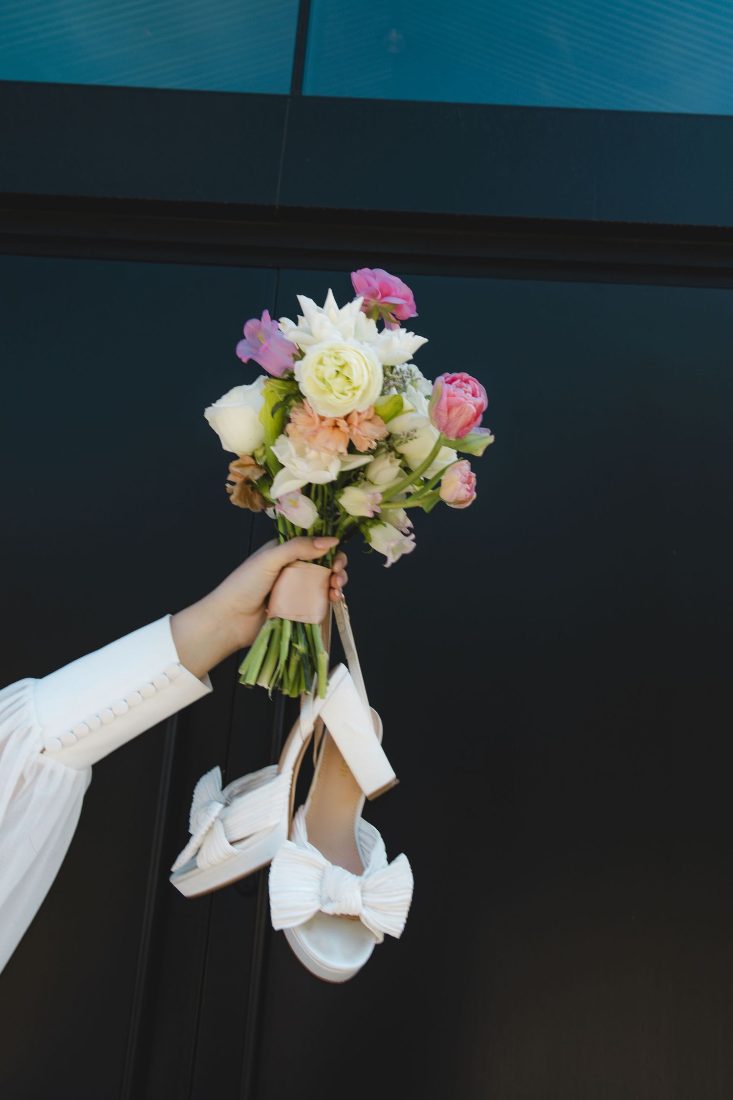 Hand holding a colorful bridal bouquet with white heels dangling against a dark background.
