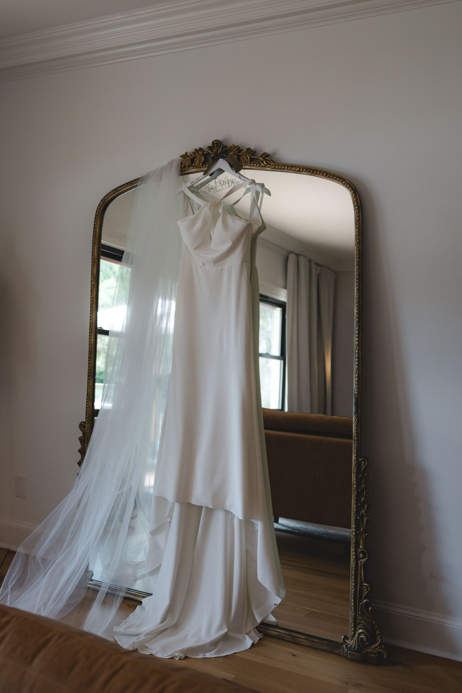 Elegant white wedding gown with veil displayed on ornate gold floor mirror in bright airy room.