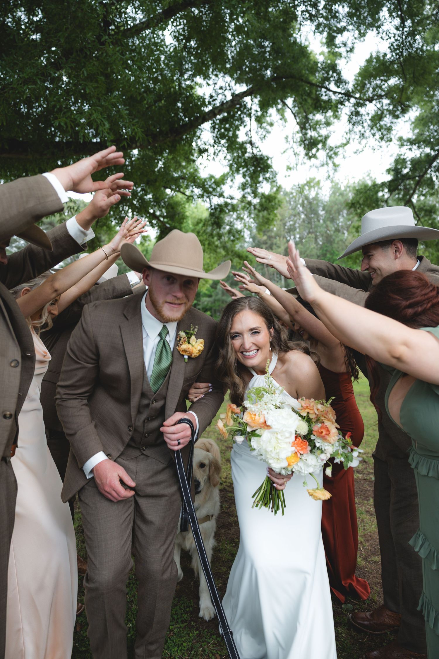 Bride and groom walk through arch of raised hands formed by wedding party in rustic outdoor ceremony.