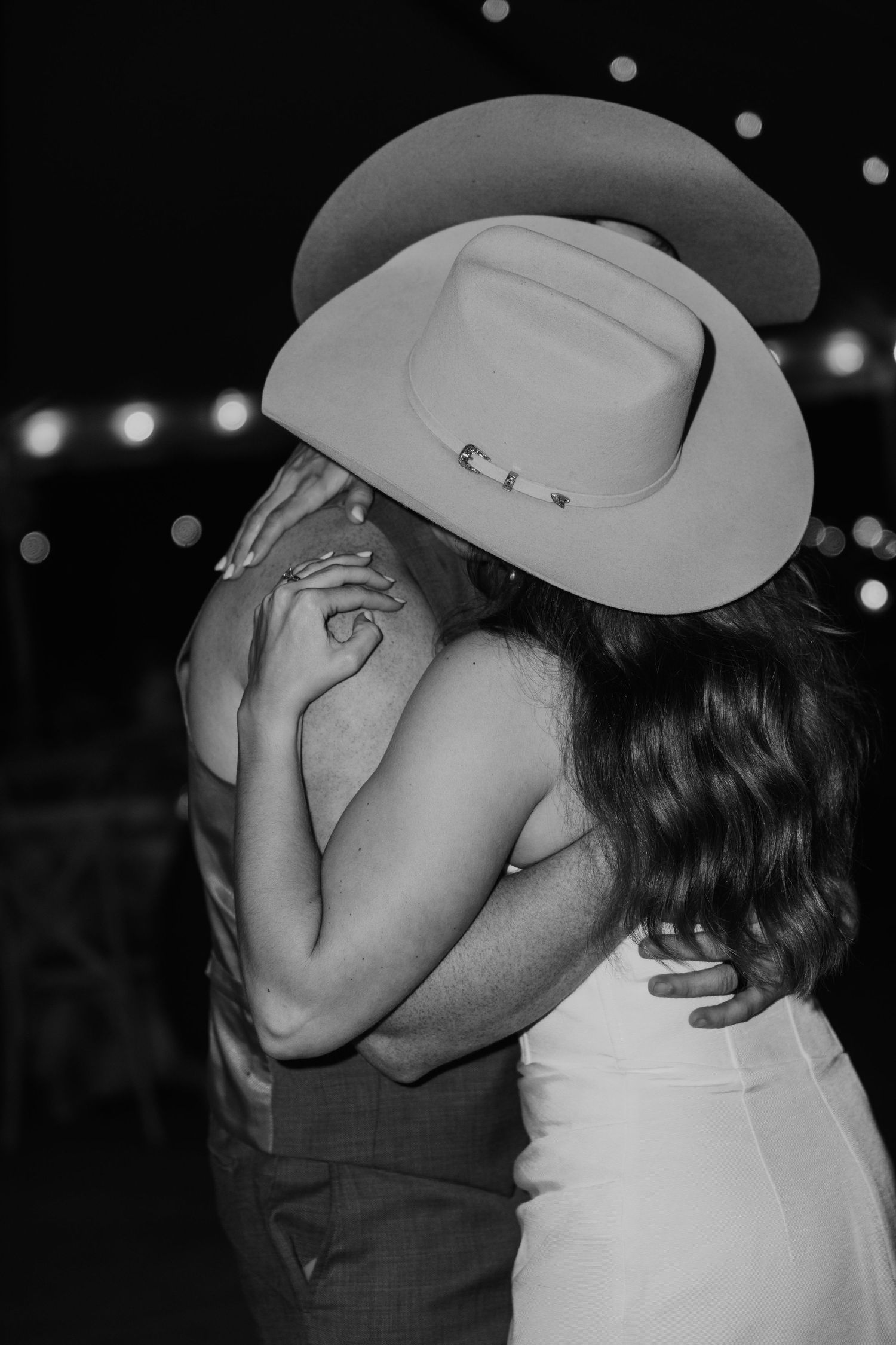 Black and white photo of bride and groom kissing under a cowboy hat at their outdoor wedding reception.