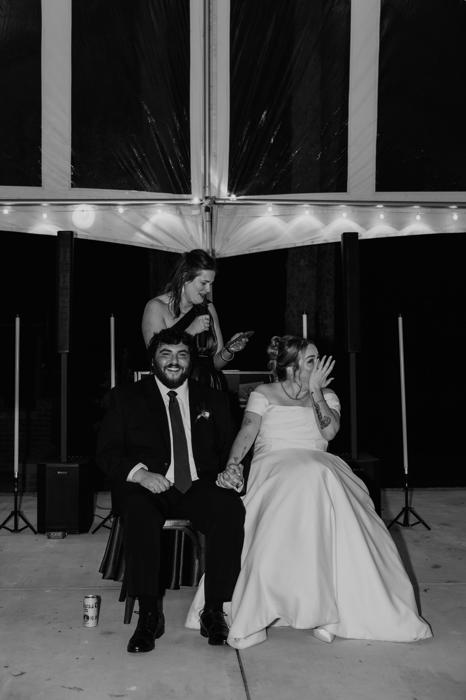 Black and white wedding photo of bride and groom seated while a bridesmaid speaks behind them at an elegant tent reception.