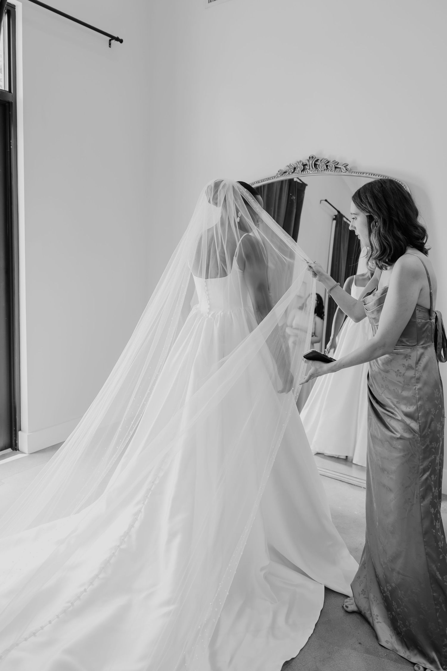 Black and white photo of a bride in a flowing gown and veil being assisted by a bridesmaid near an ornate mirror.