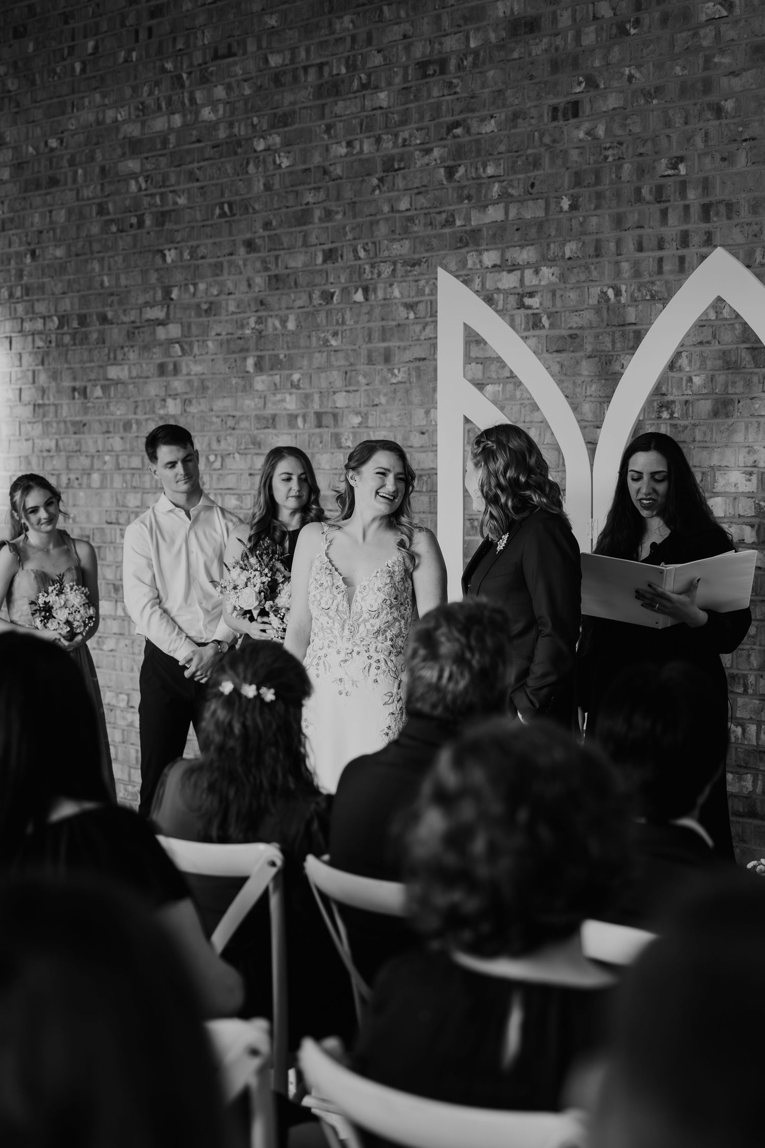 Black and white wedding ceremony photo with bride, groom, and officiant at brick wall venue.