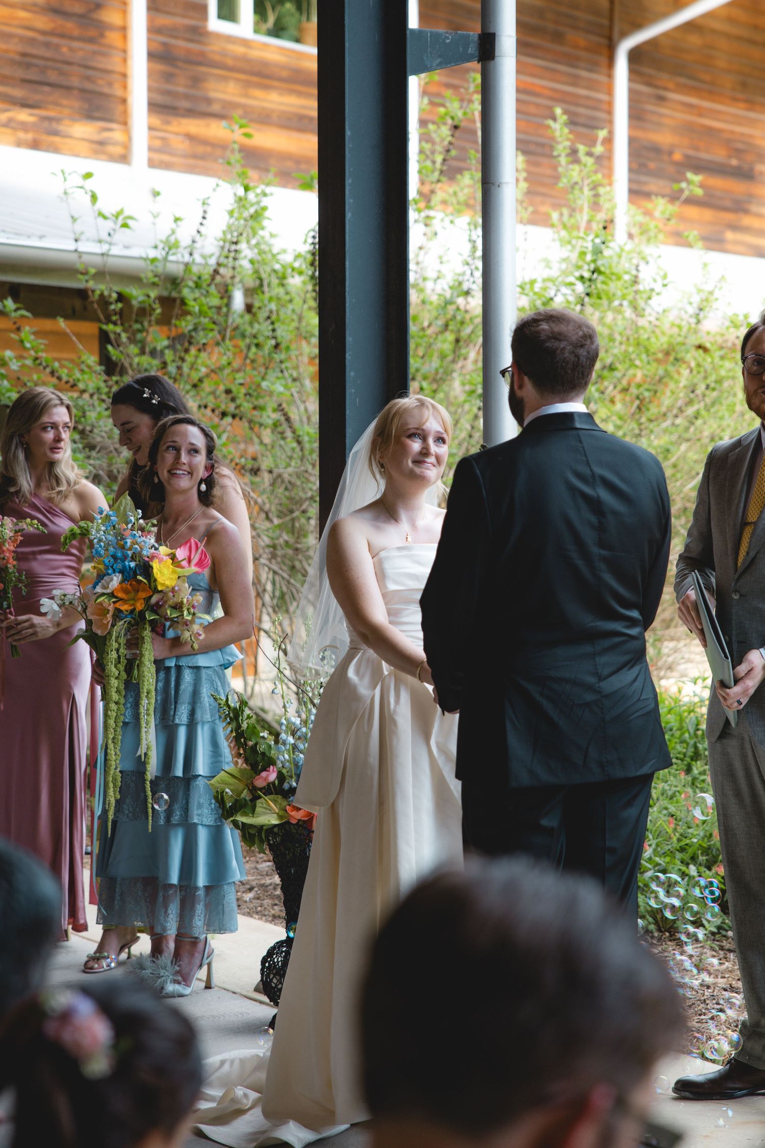 Smiling bride in white gown exchanges vows with groom at outdoor wedding ceremony with bridesmaids holding colorful bouquets.