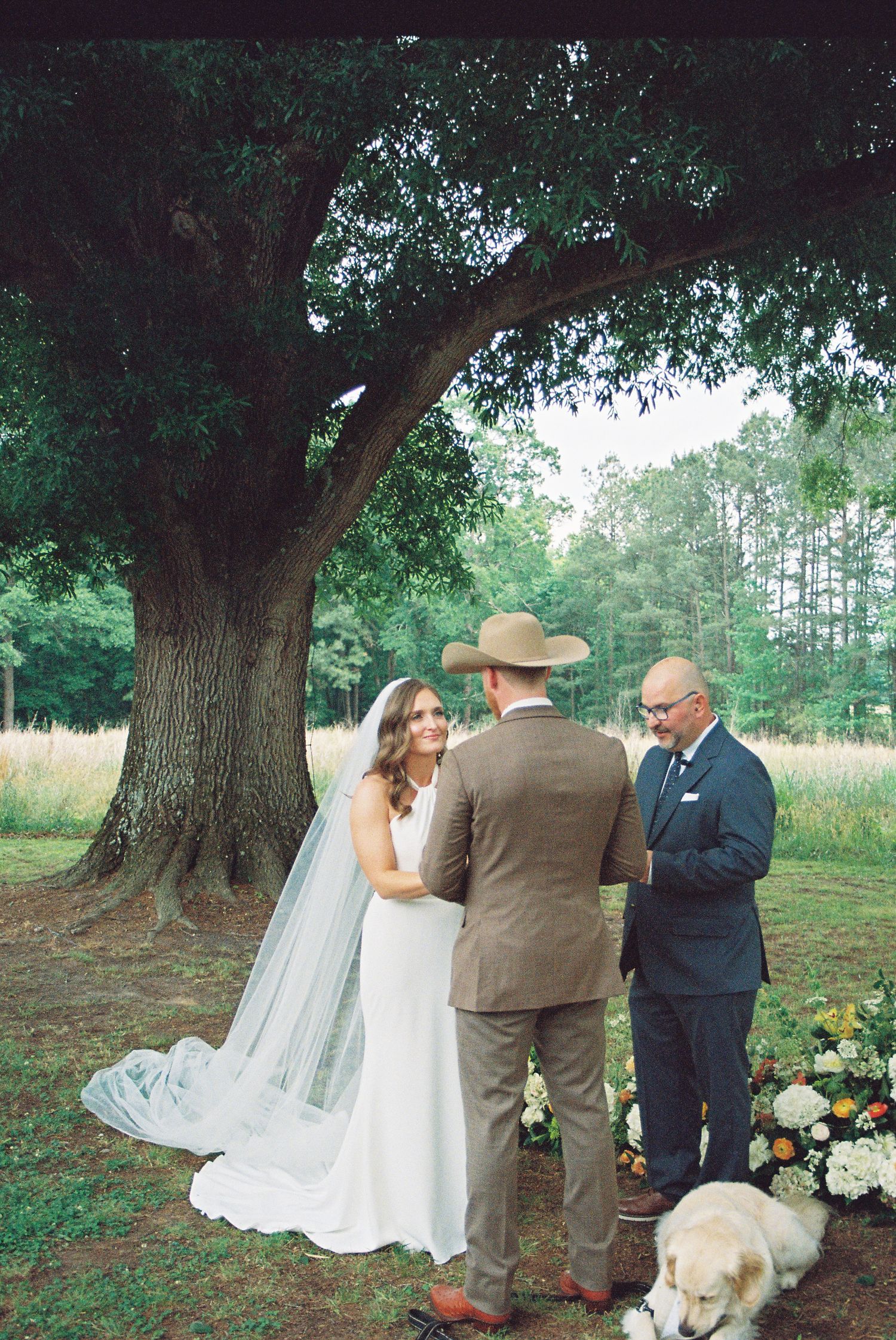 Outdoor wedding ceremony under large oak tree with bride in white gown and veil, groom in brown suit, officiant, and dog.