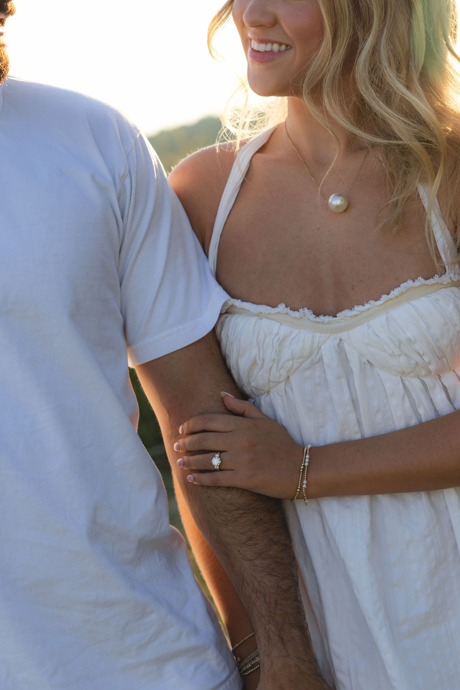Smiling blonde woman in white dress with pearl necklace and ring holds man's arm outdoors in golden light.