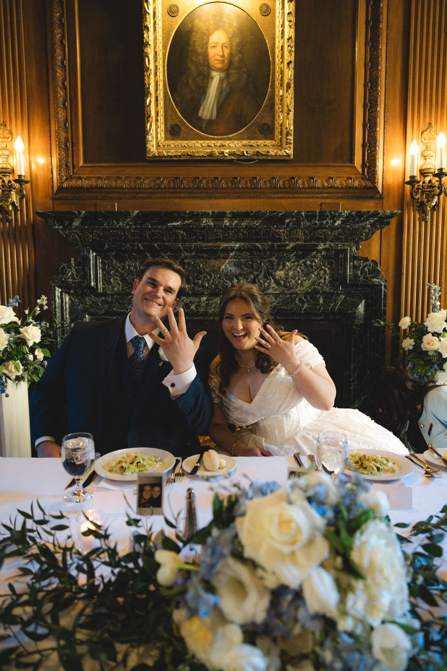 Newlyweds show off wedding rings at elegant reception table with floral arrangements and ornate fireplace backdrop.