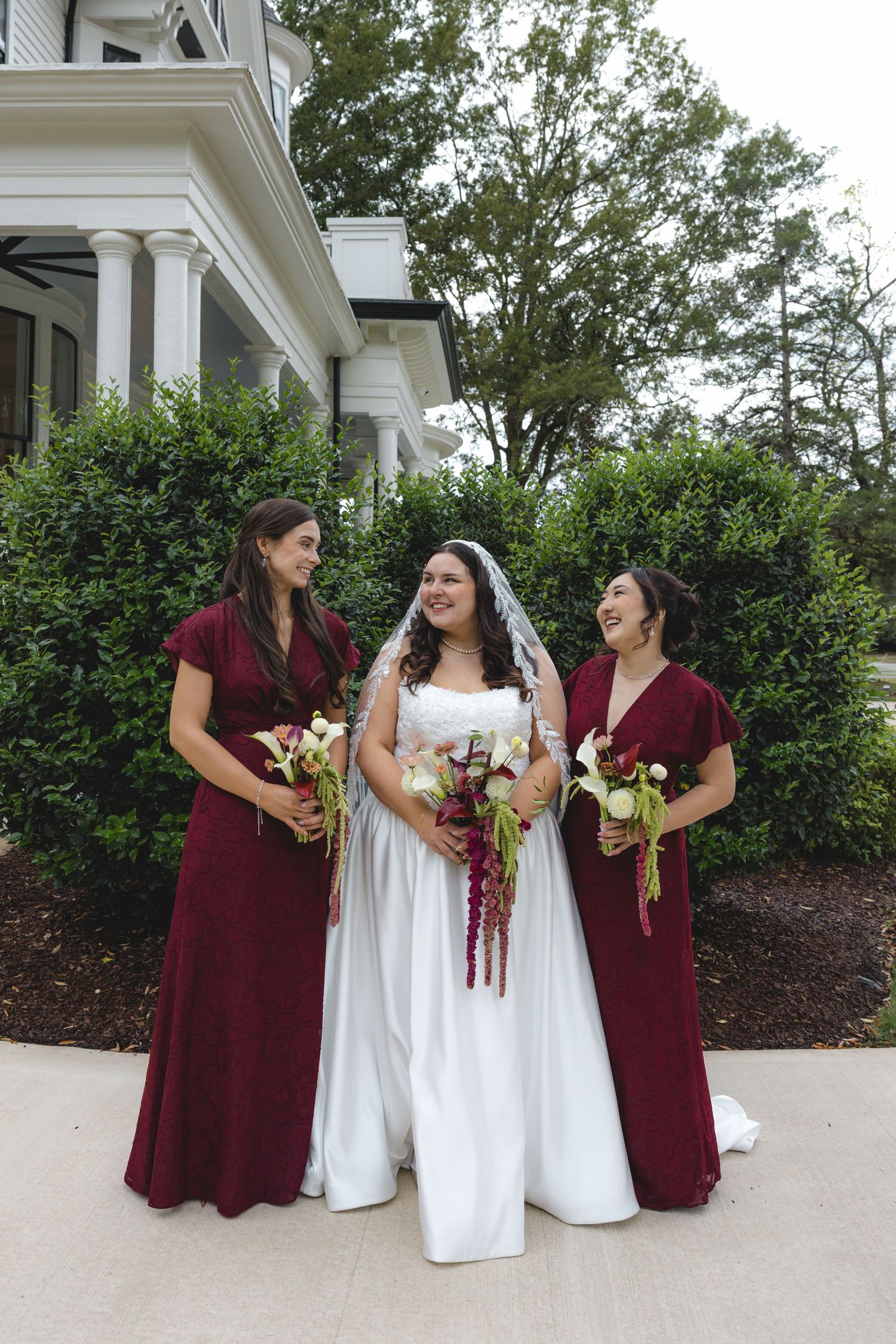 A bride in a white gown and veil laughs with two bridesmaids in burgundy dresses, all holding bouquets outside a historic white building.