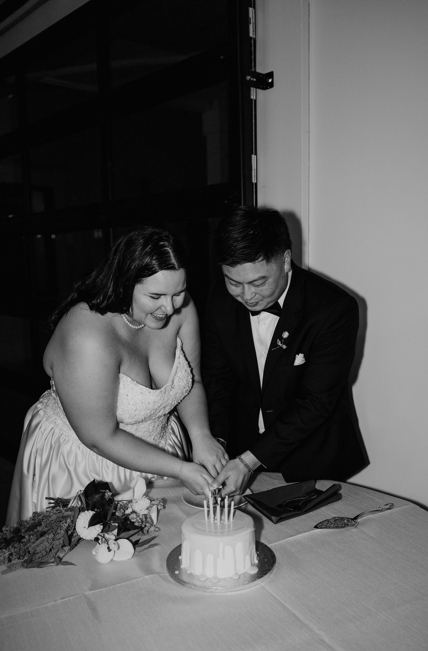 Black and white photo of a bride and groom smiling as they cut their small wedding cake with candles, with a floral bouquet beside them.