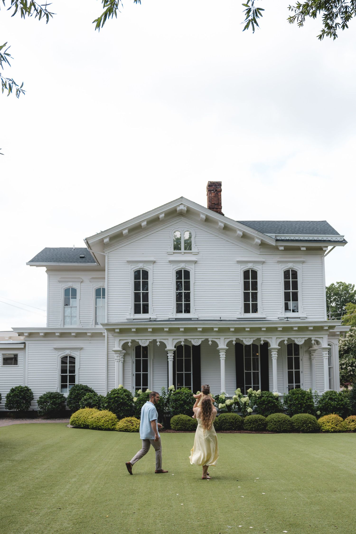 Couple walking toward a grand white two-story colonial home with lush green lawn and manicured shrubs.