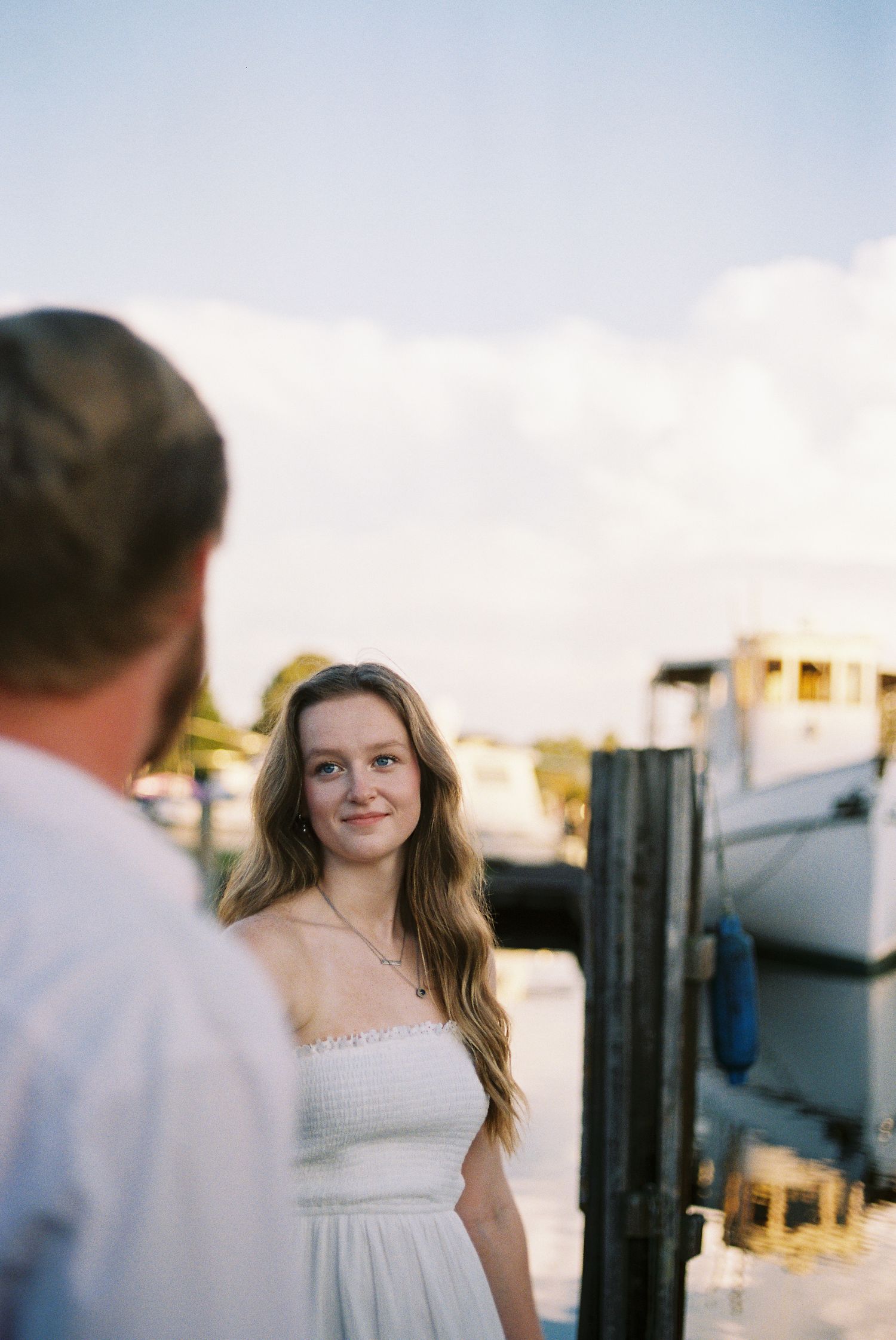 A young woman in a white dress smiles at a man near a marina dock with boats in the background.