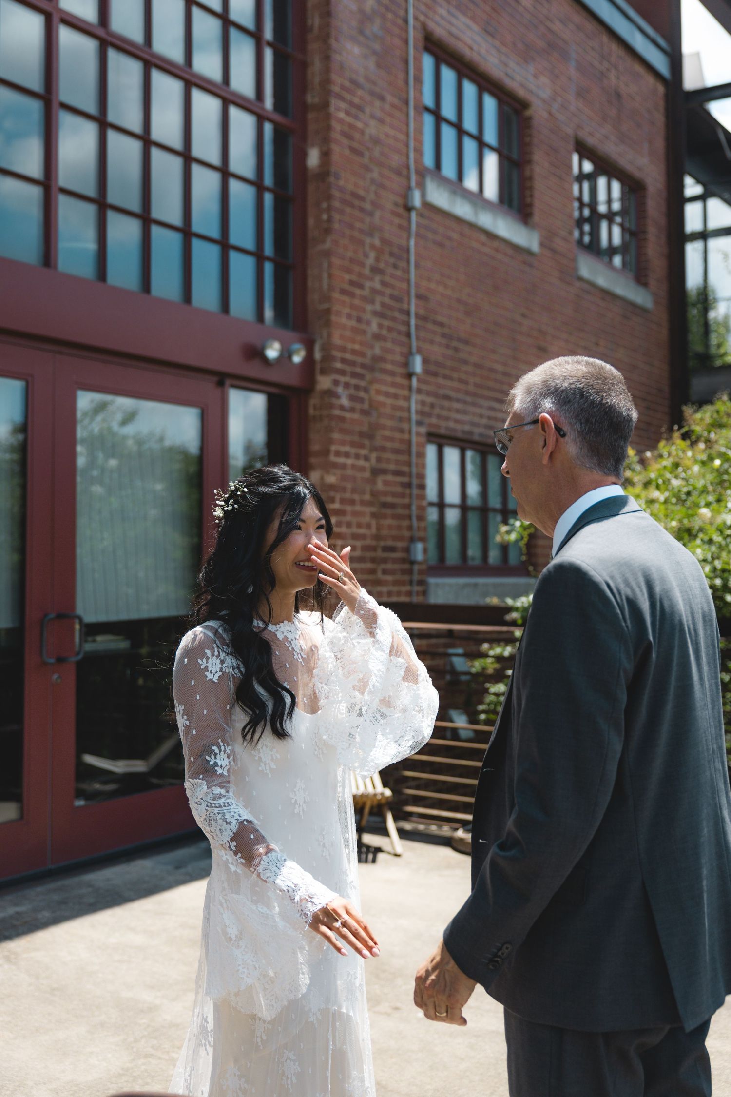 Bride in lace gown covers mouth in emotional reaction during first look with groom outside brick venue.