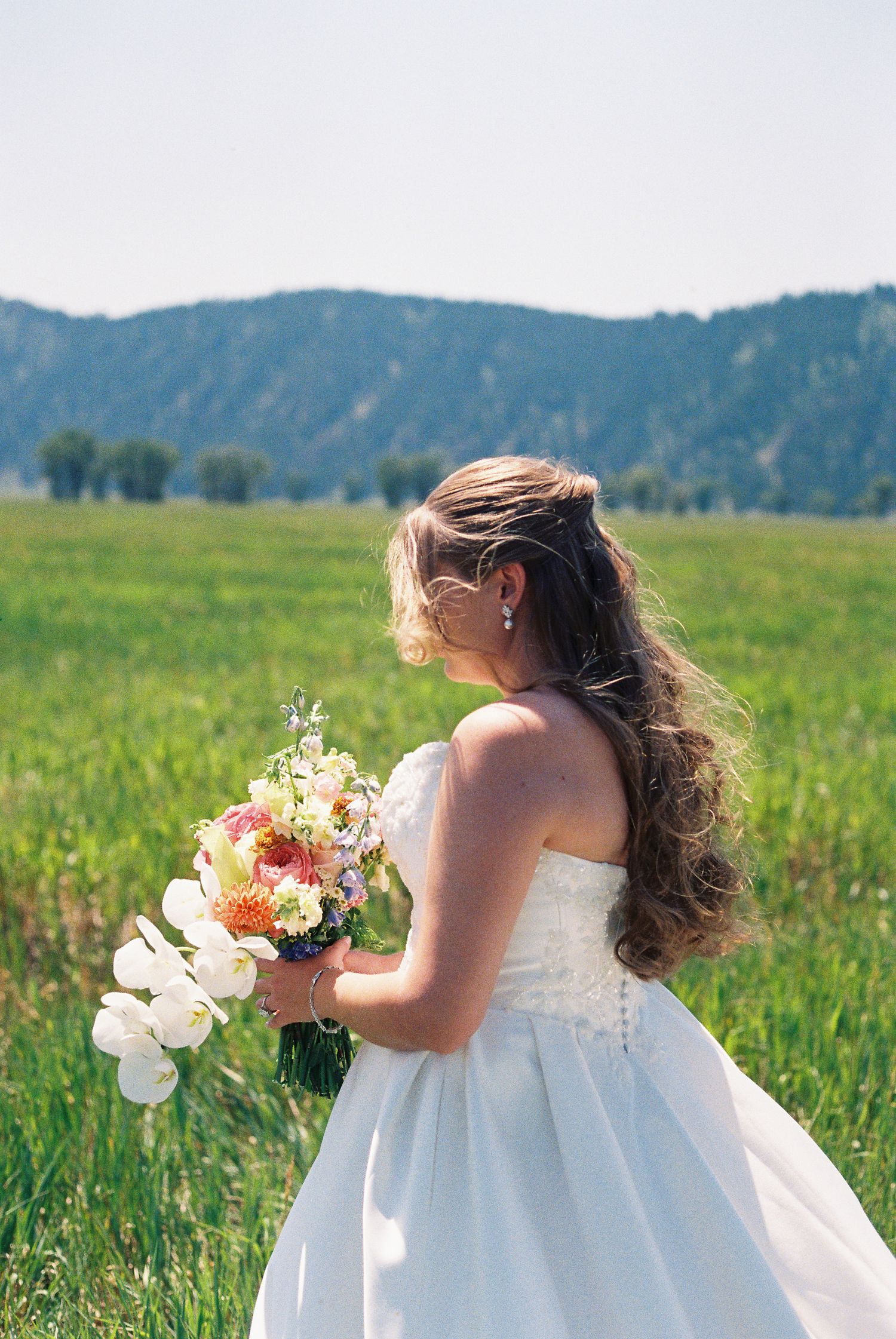 Bride in white strapless gown holding floral bouquet standing in green meadow with mountain backdrop on sunny day.