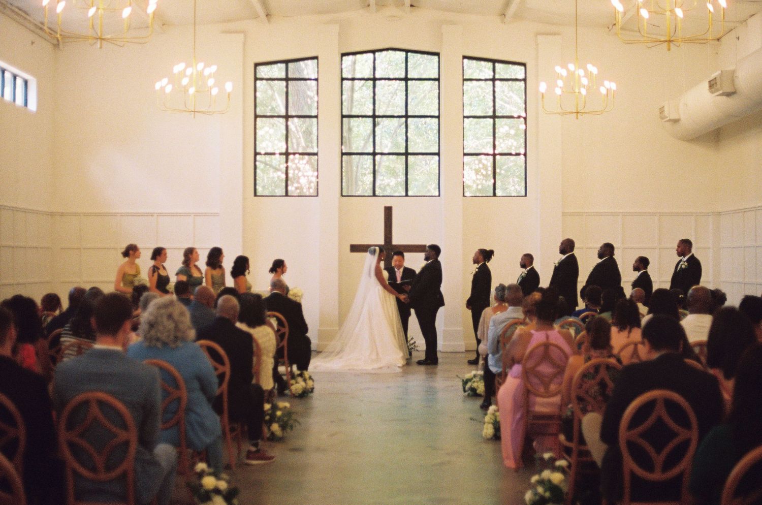 Elegant wedding ceremony in a white chapel with cross, bride and groom at altar, guests seated in wooden pews.